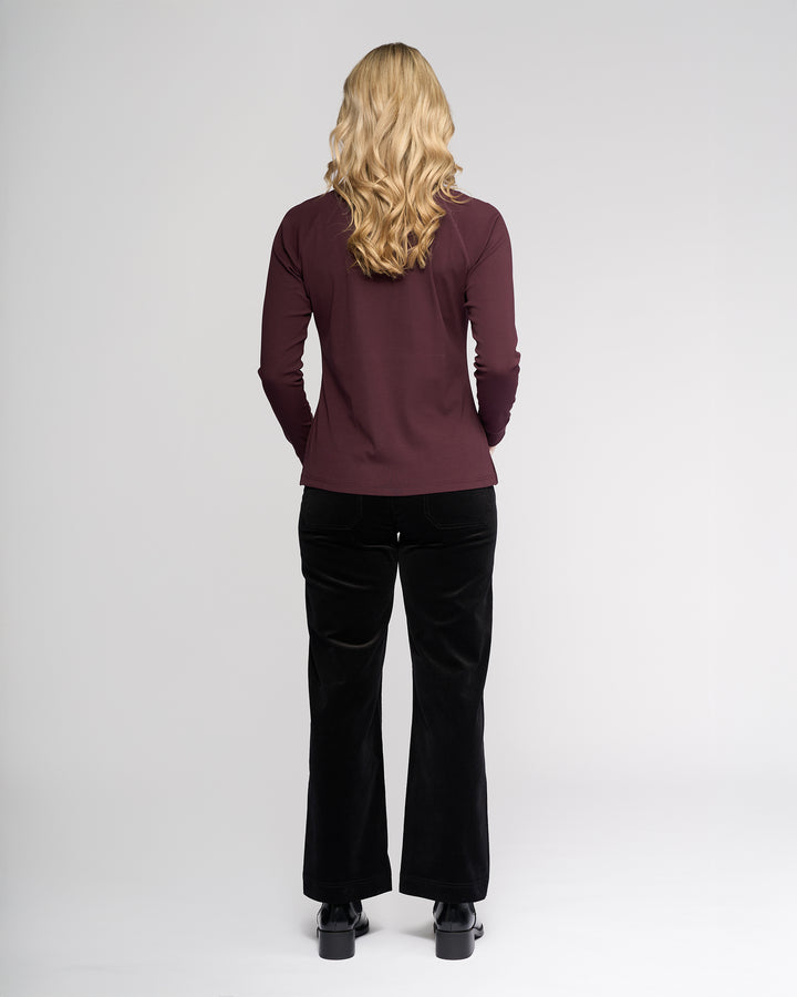 A person with long, wavy blonde hair stands with their back to the camera, wearing the Vassalli Key Top - Pinot paired with black pants and black shoes against a plain light backdrop for a casual look.