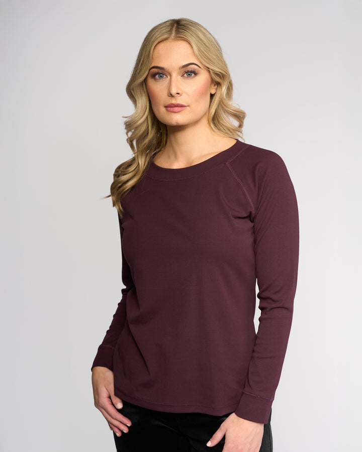 A woman with long blonde hair wears the Vassalli Key Top - Pinot, a maroon stretch cotton long-sleeve top, paired with black pants. She stands against a plain light gray background, looking forward with a neutral expression for a casual look.