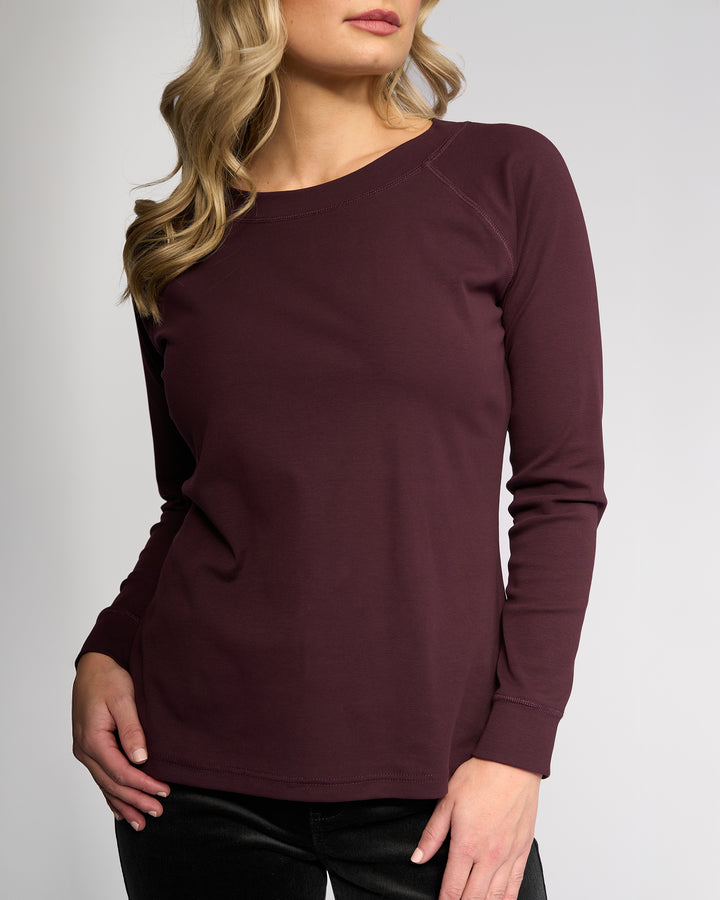 A woman with wavy blonde hair wears the Vassalli Key Top - Pinot, a casual long-sleeve stretch cotton burgundy top, paired with black pants. She stands against a plain light background, her head slightly turned and left hand on her hip.