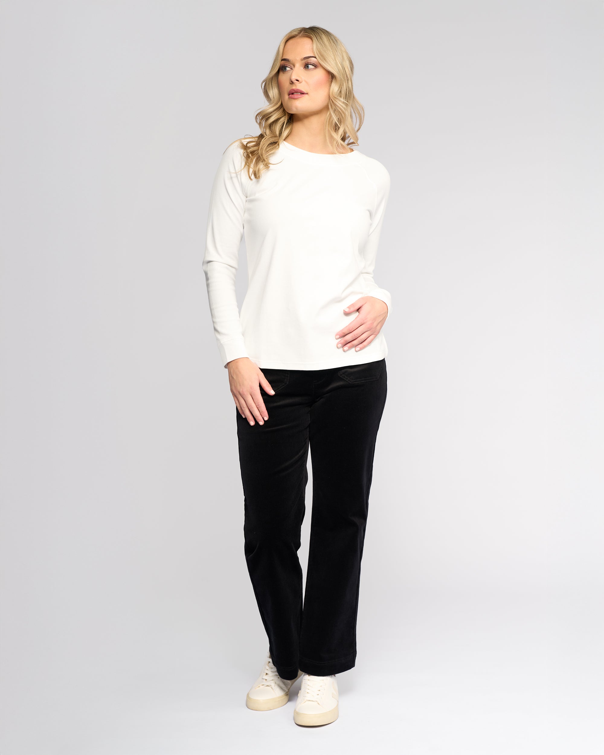 A woman with long blonde hair wears the Vassalli Key Top in Winter White, styled casually with black pants and white sneakers, standing against a light background and looking left with one hand on her hip.