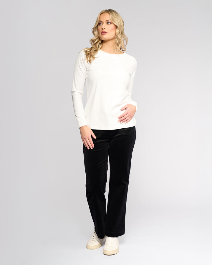 A woman with long blonde hair wears the Vassalli Key Top in Winter White, styled casually with black pants and white sneakers, standing against a light background and looking left with one hand on her hip.