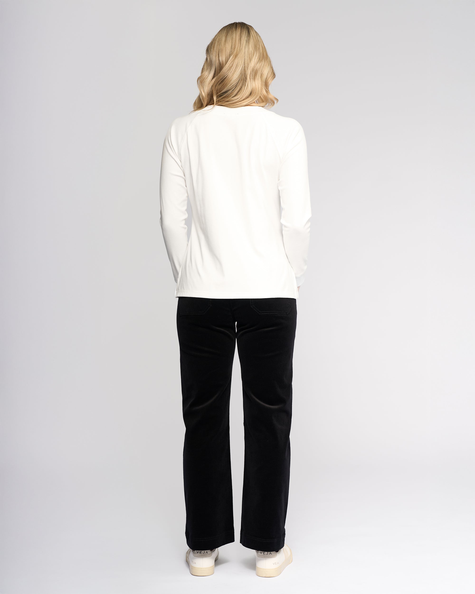 A woman with blonde hair faces away from the camera, wearing the Vassalli Key Top in Winter White with black pants and white shoes against a plain light background—an ideal choice for a casual everyday look.