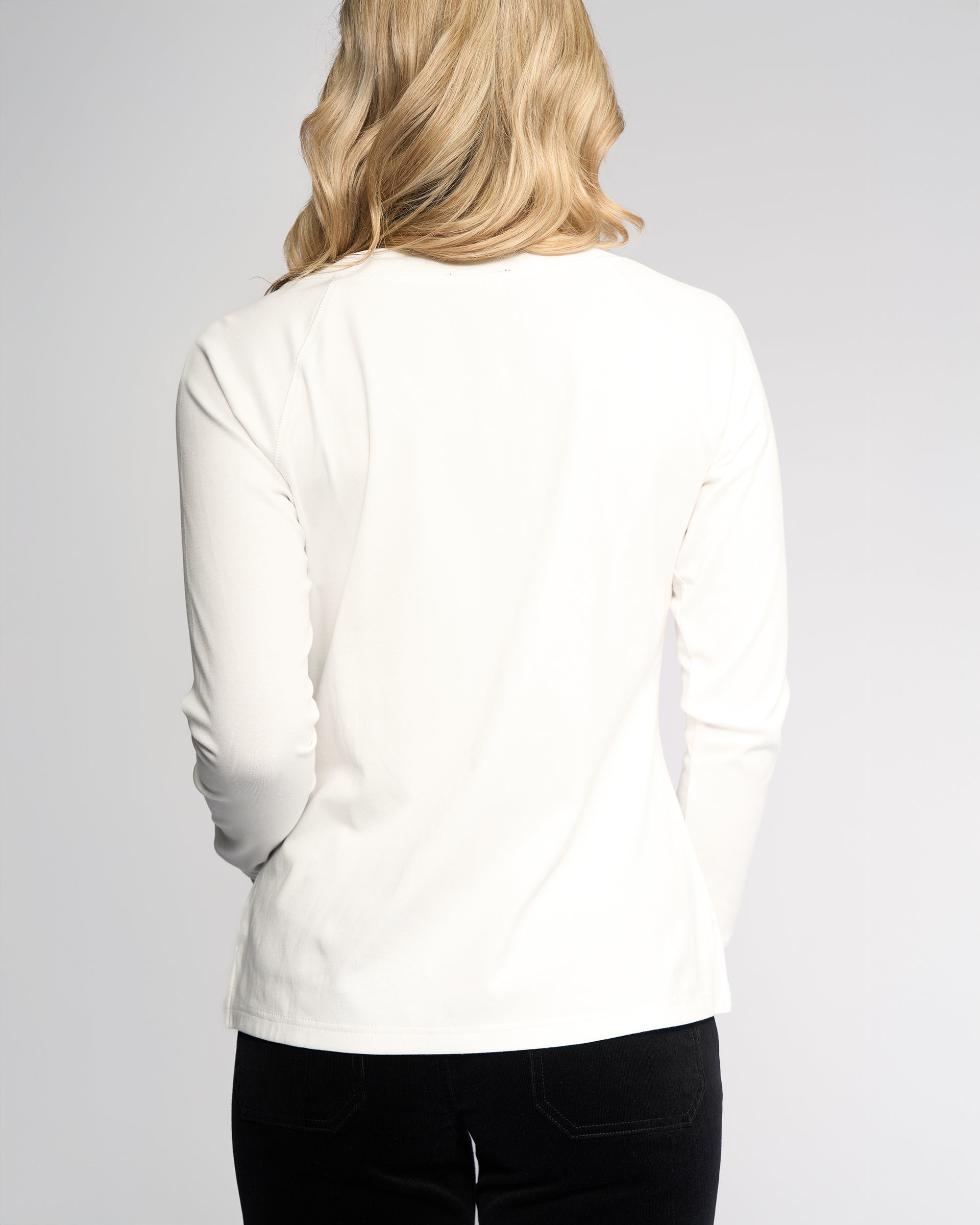 A person with long blonde hair is shown from the back, wearing the Vassalli Key Top in Winter White paired with black pants, set against a plain light background.
