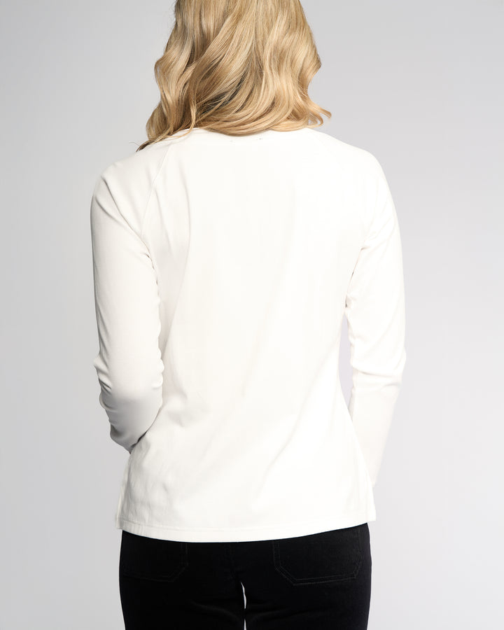 A person with long blonde hair is shown from the back, wearing the Vassalli Key Top in Winter White paired with black pants, set against a plain light background.