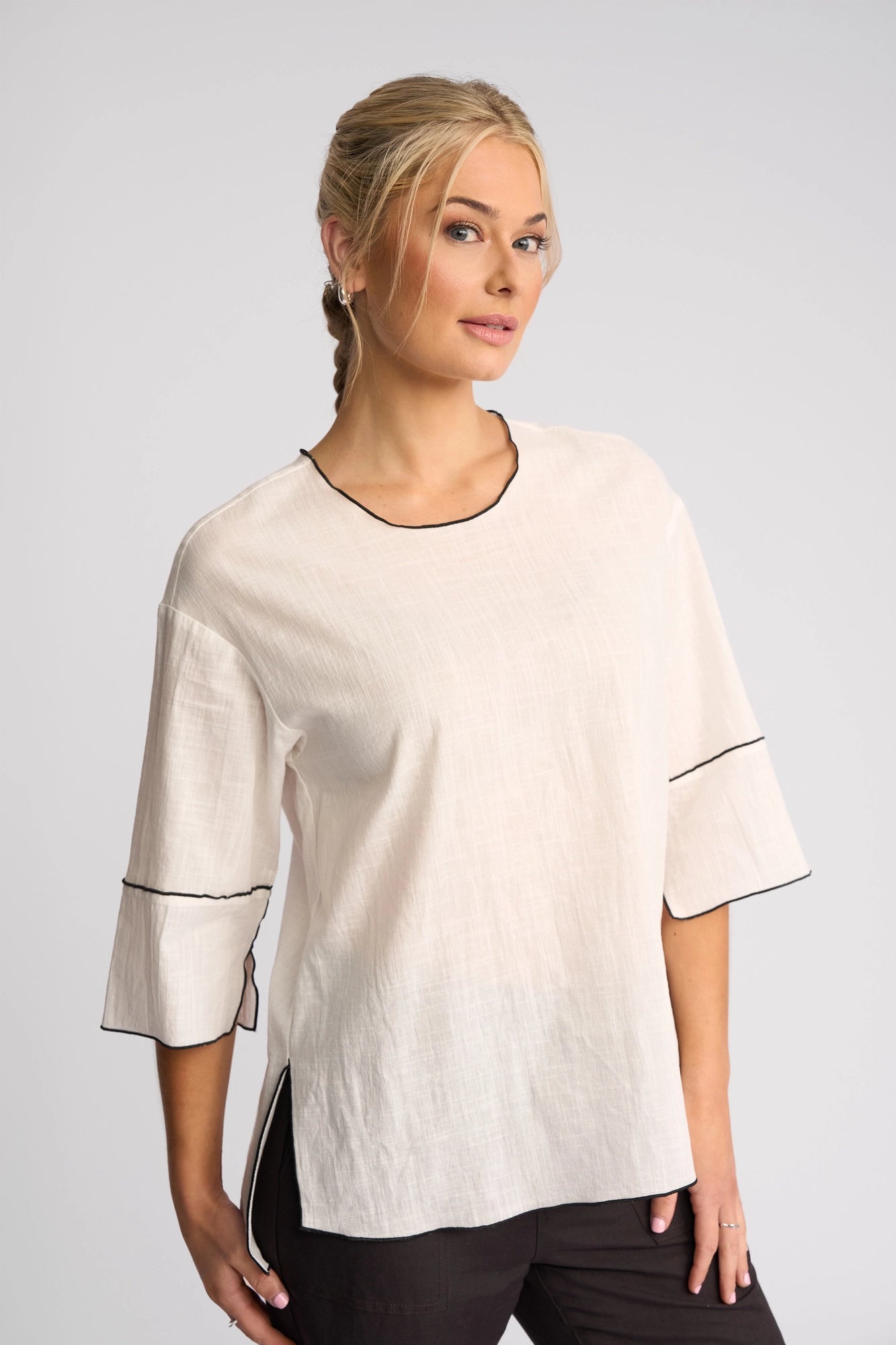 A woman with blonde hair in a braid wears the Vassalli Sloane Top in Off White, featuring black trim, paired with dark pants. She stands against a plain light gray background, looking slightly to the side.