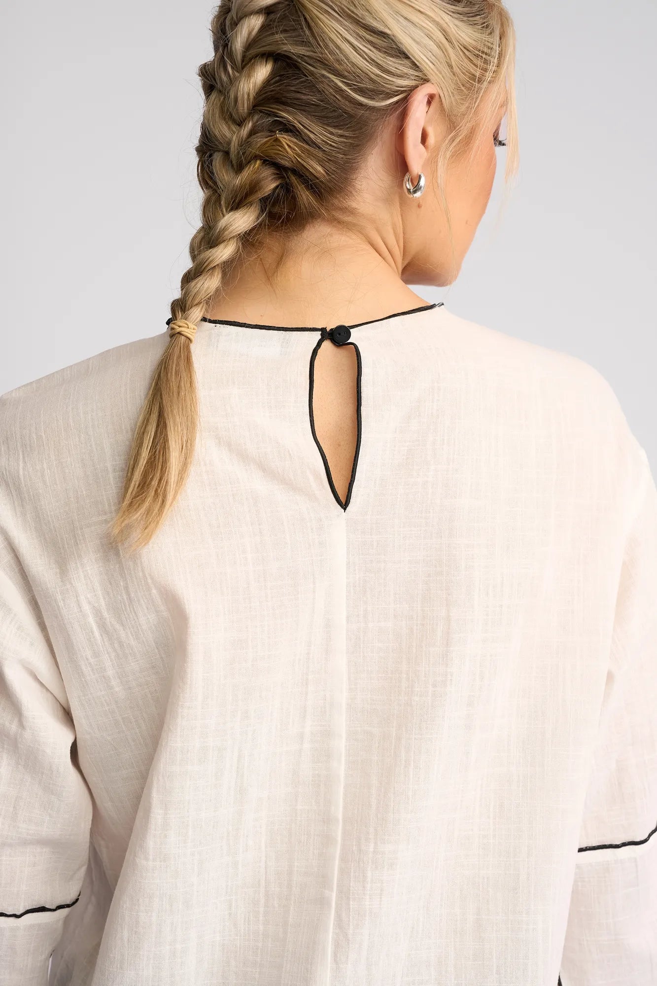 A woman with blonde hair in a braid is shown from behind, wearing the Vassalli Sloane Top in Off White, featuring black trim and a button closure at the back of the neck.