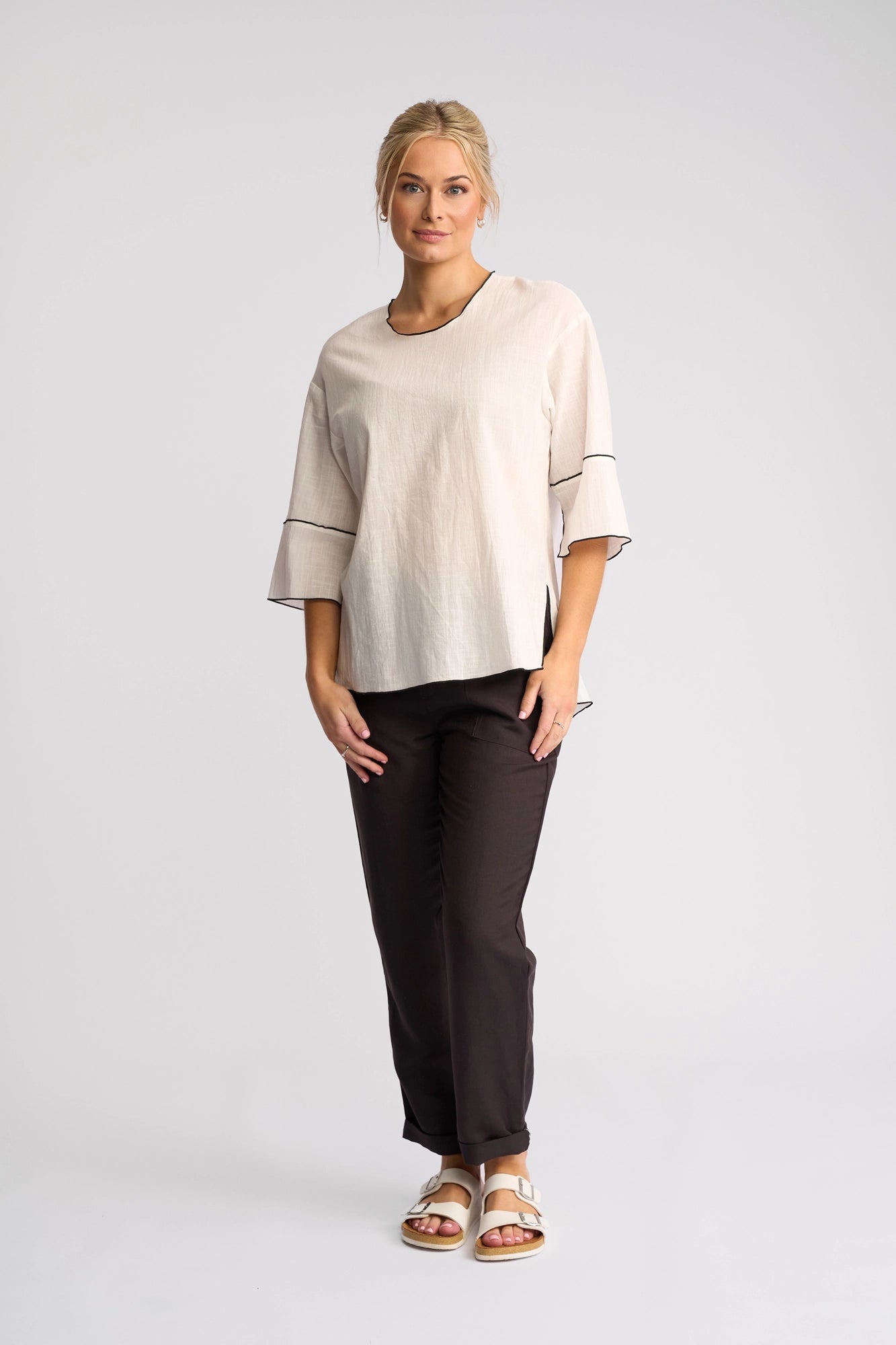 A woman stands against a plain background, wearing the Vassalli Sloane Top in Off White, a lightweight cotton piece with three-quarter sleeves. She pairs it with dark pants and white sandals. Her blonde hair is pulled back, and she looks forward with a slight smile.