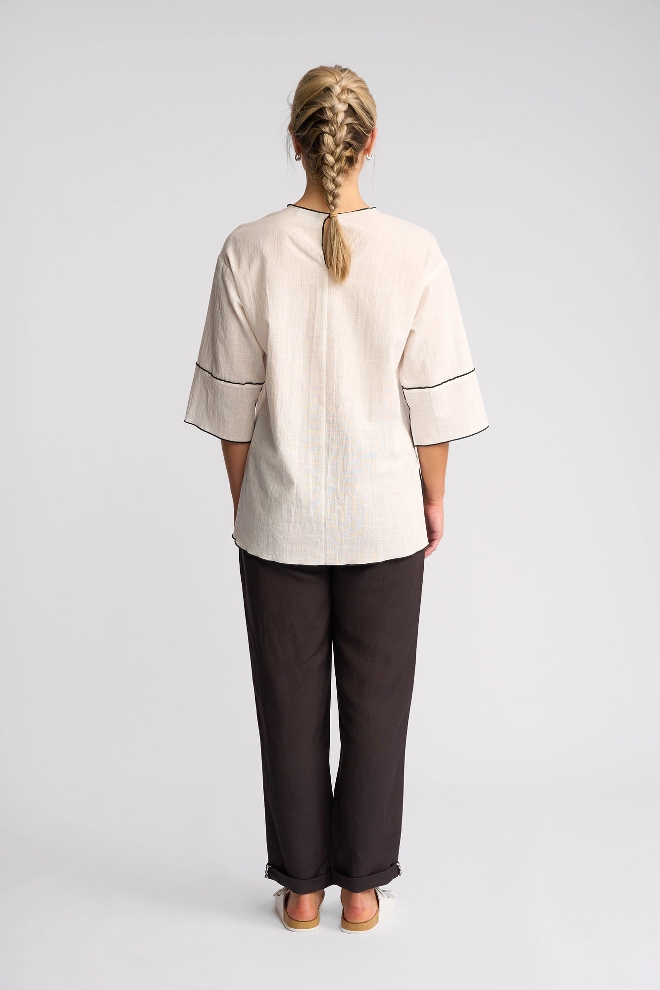 A person with blonde braided hair stands facing away, wearing the Vassalli Sloane Top in Off White—a loose, oversized style with wide sleeves and black trim—paired with black pants and light-colored shoes against a plain white background.