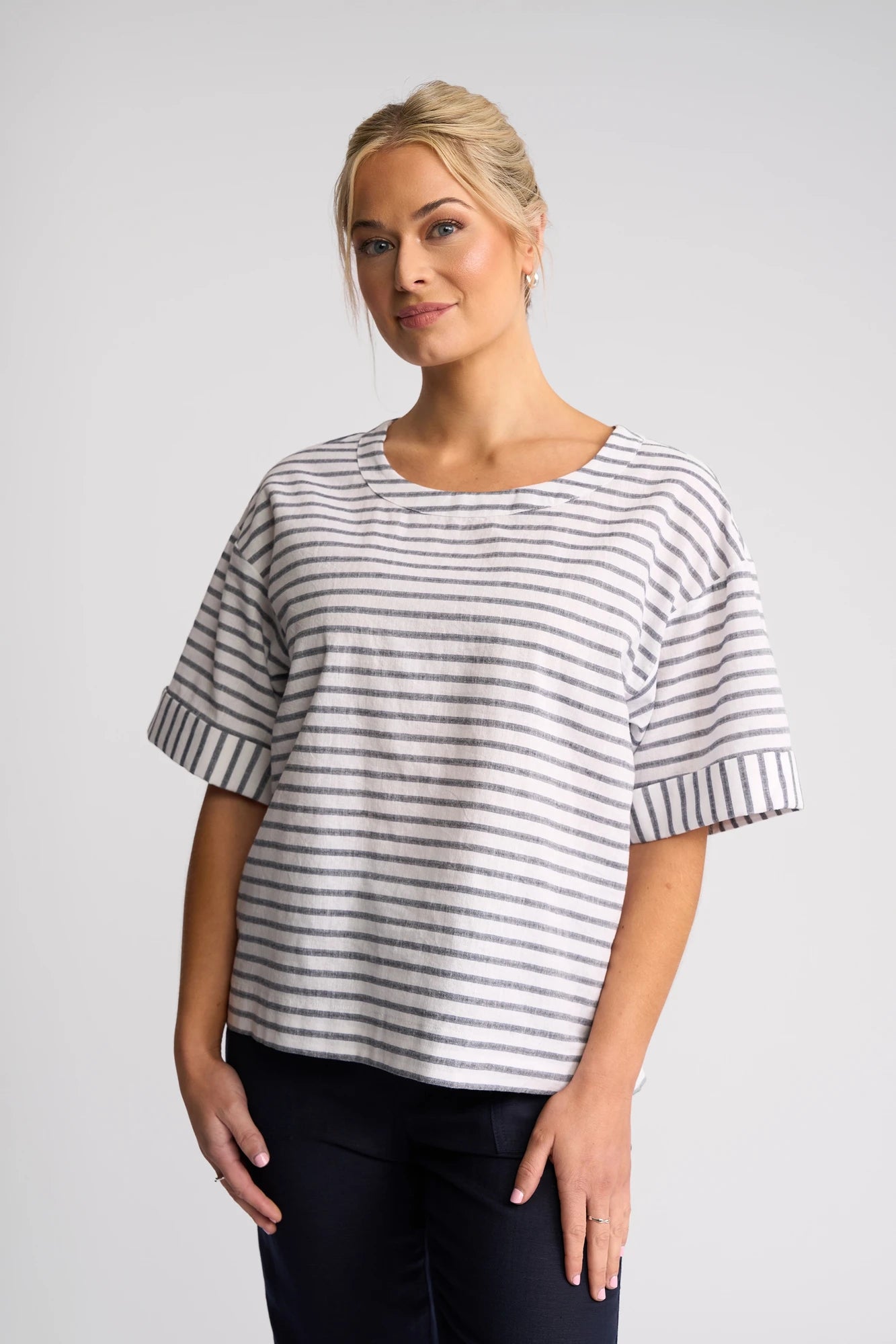 A woman with blonde hair in a loose bun stands against a plain light background, softly smiling with her hands at her sides. She is wearing the Vassalli Optic Top in Black Stripe—a relaxed cotton-linen blend shirt with grey and white stripes—paired with dark pants.