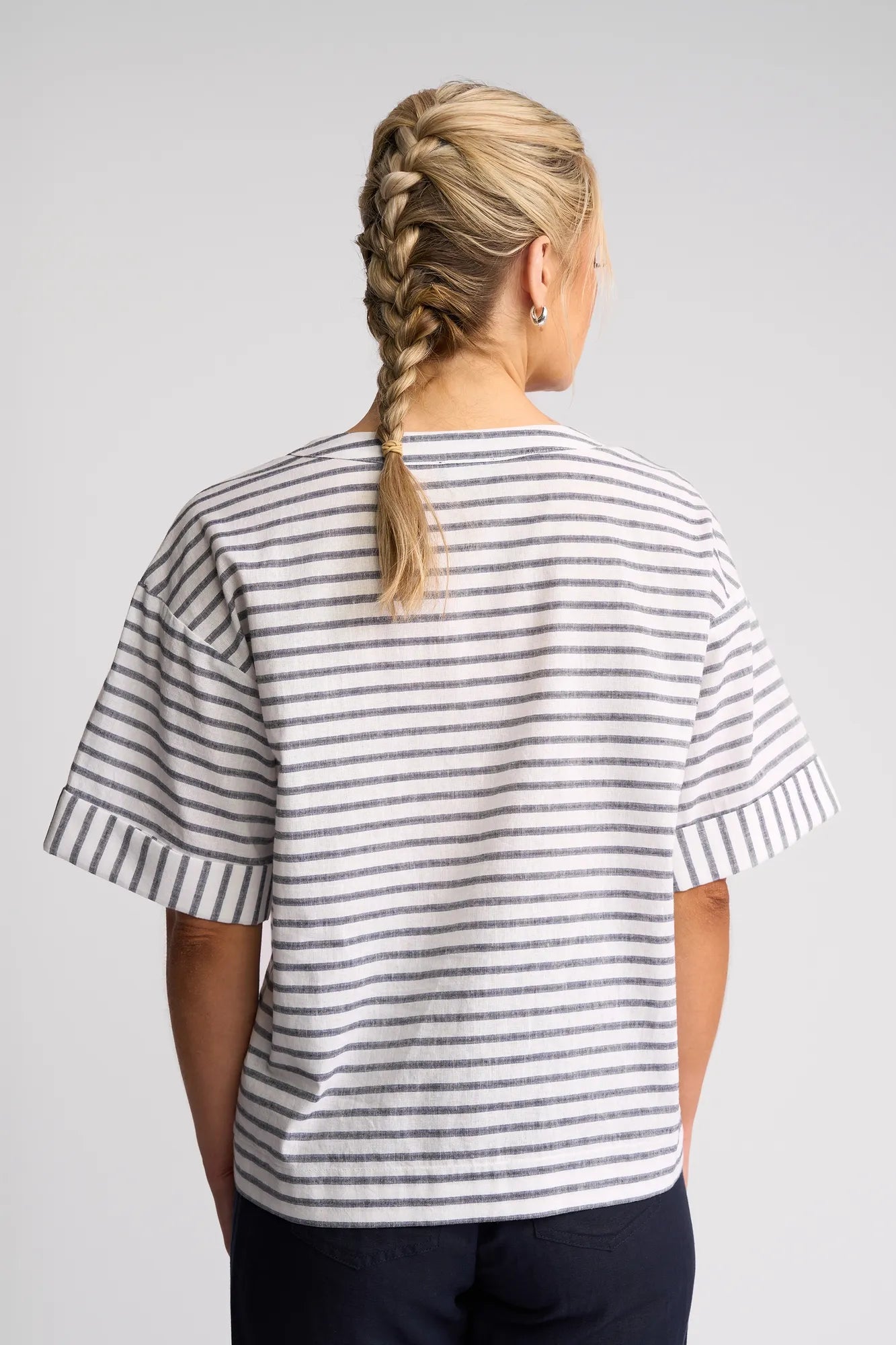 A person with blonde hair in a braid is seen from the back, wearing the Vassalli Optic Top - Black Stripe—a relaxed cotton-linen blend top with grey and white stripes—and dark pants, standing against a plain light background.