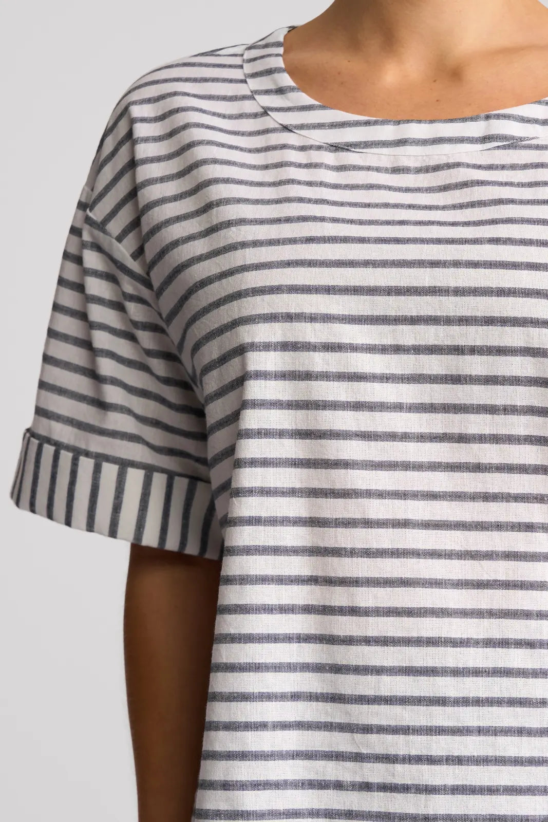 A person wearing the Vassalli Optic Top - Black Stripe, a relaxed short-sleeved shirt featuring horizontal black stripes on a white background. Crafted from a comfortable cotton-linen blend, the top has a round neckline and cuffed sleeves. Only the upper torso is visible.