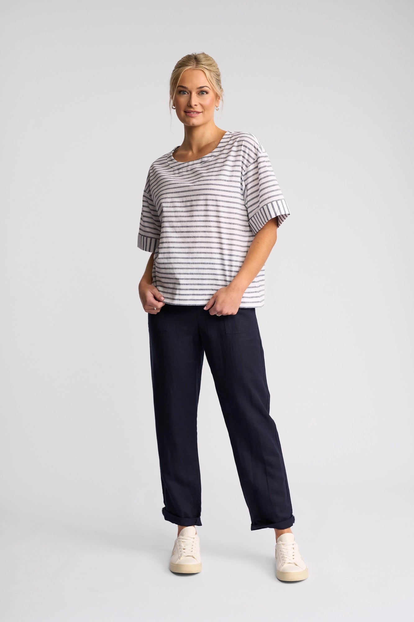 A woman stands facing forward wearing the Vassalli Optic Top in Black Stripe, a relaxed short-sleeved cotton-linen blend, styled with dark pants and white sneakers. Her blonde hair is pulled back as she poses against a plain light background.