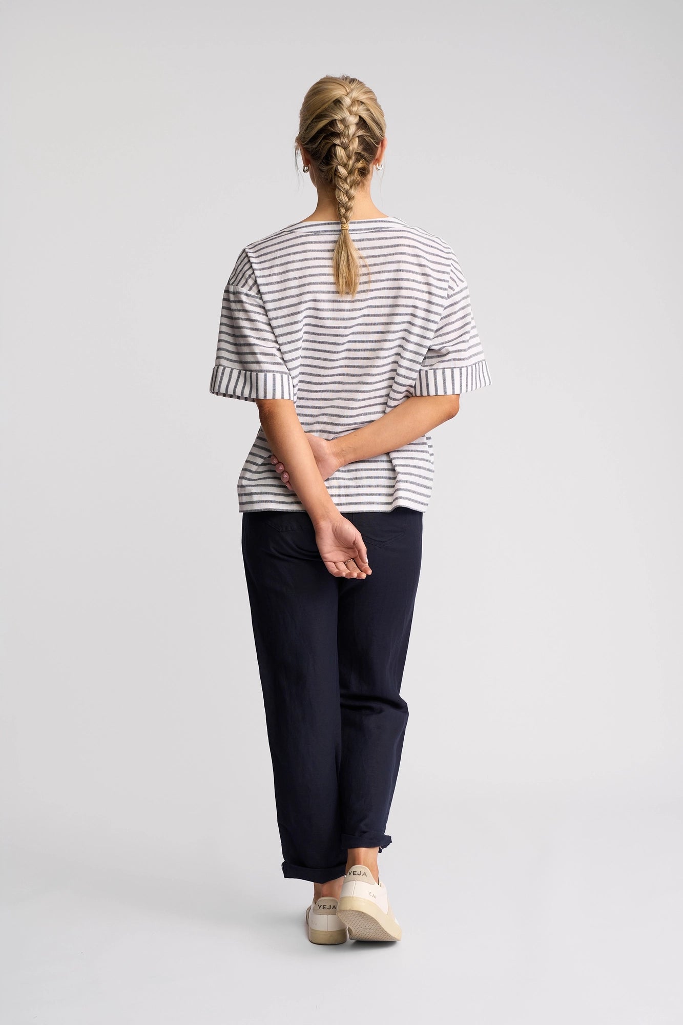 A person with blonde hair in a braid stands facing away, wearing the Vassalli Optic Top in Black Stripe—a relaxed cotton-linen blend top—paired with dark pants and light sneakers, arms crossed behind their back against a plain background.