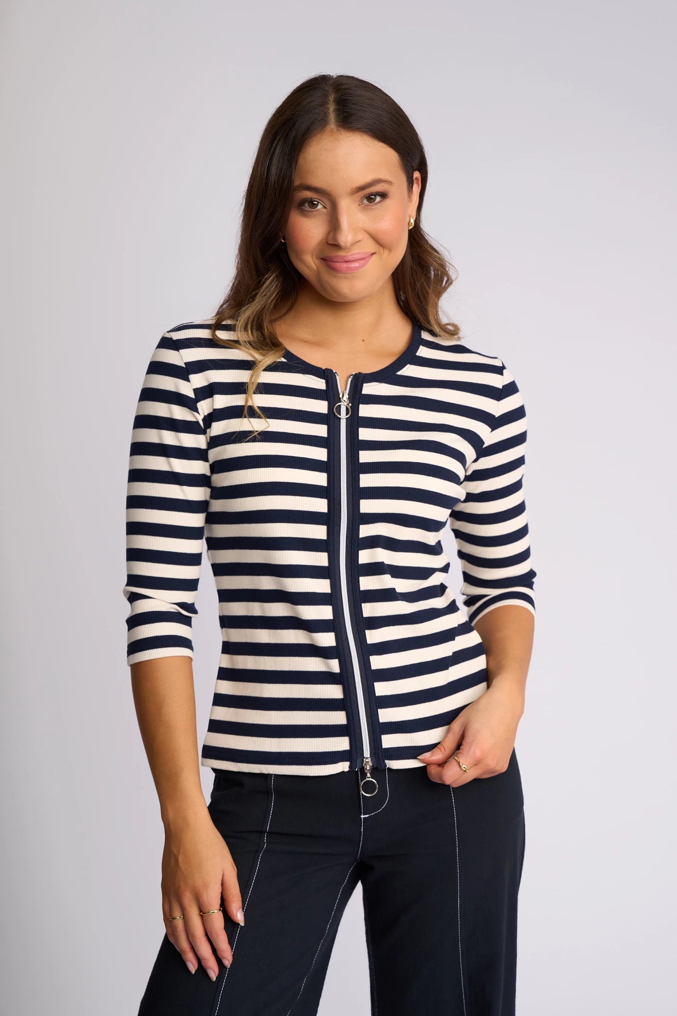 A woman smiles while standing against a plain light background, wearing the Vassalli Callie Top in Ink/Cream—a ribbed knit top featuring chic ink and cream stripes with a stylish two-way zip front—paired with dark pants.