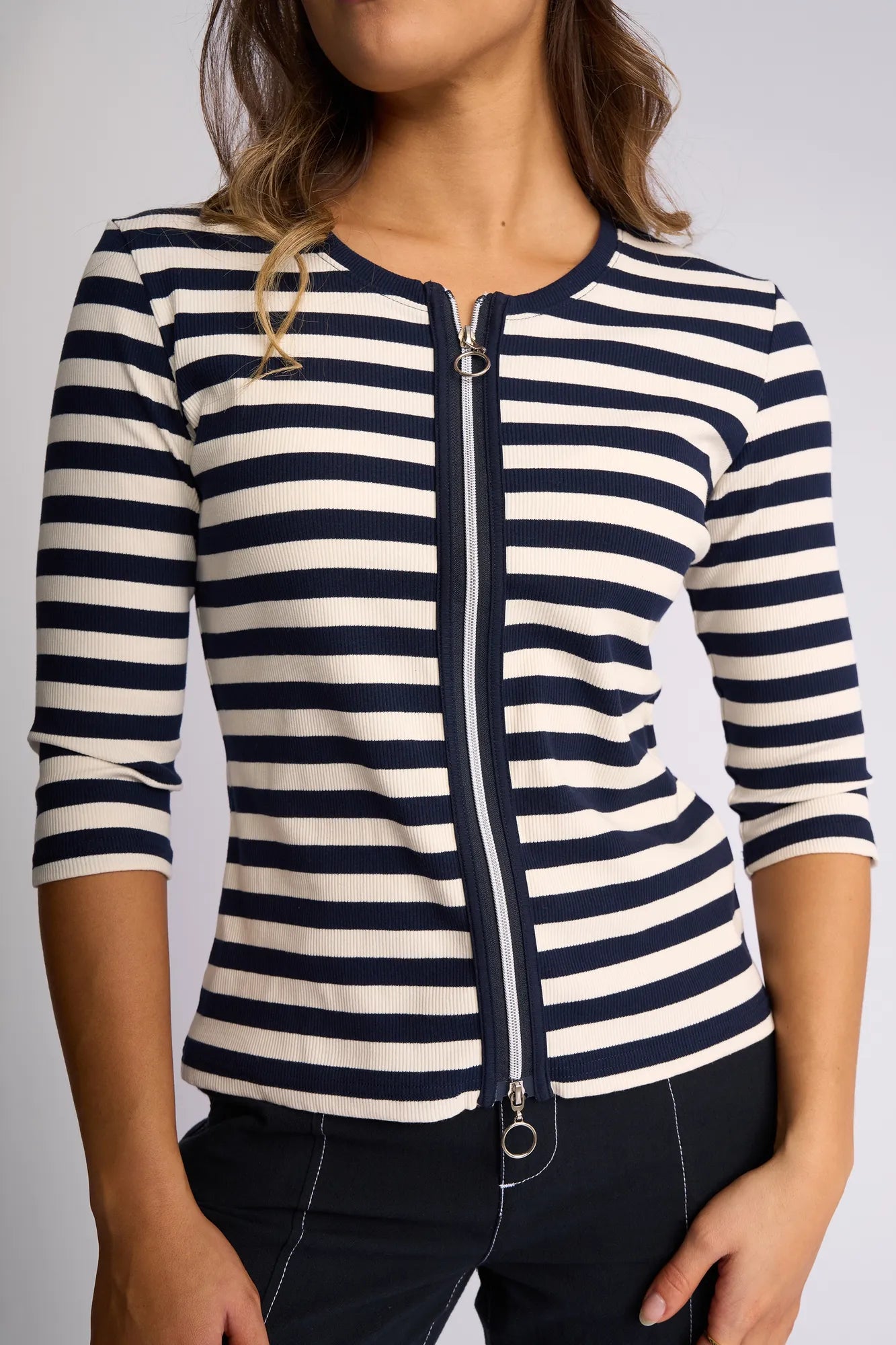 A woman stands against a plain light background wearing the Vassalli Callie Top in Ink/Cream, a ribbed knit top with ink and cream horizontal stripes, featuring three-quarter sleeves and a practical two-way zip front for versatile styling. She pairs it with dark pants.