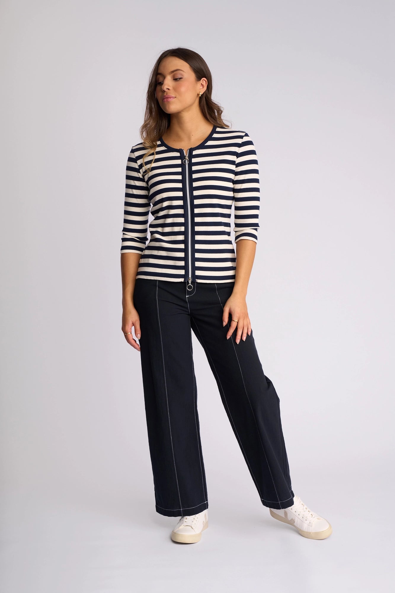 A woman stands against a plain background, wearing the Vassalli Callie Top in Ink/Cream—a ribbed knit striped top with a two-way zip front—paired with wide-leg navy pants featuring white stitching and white sneakers.