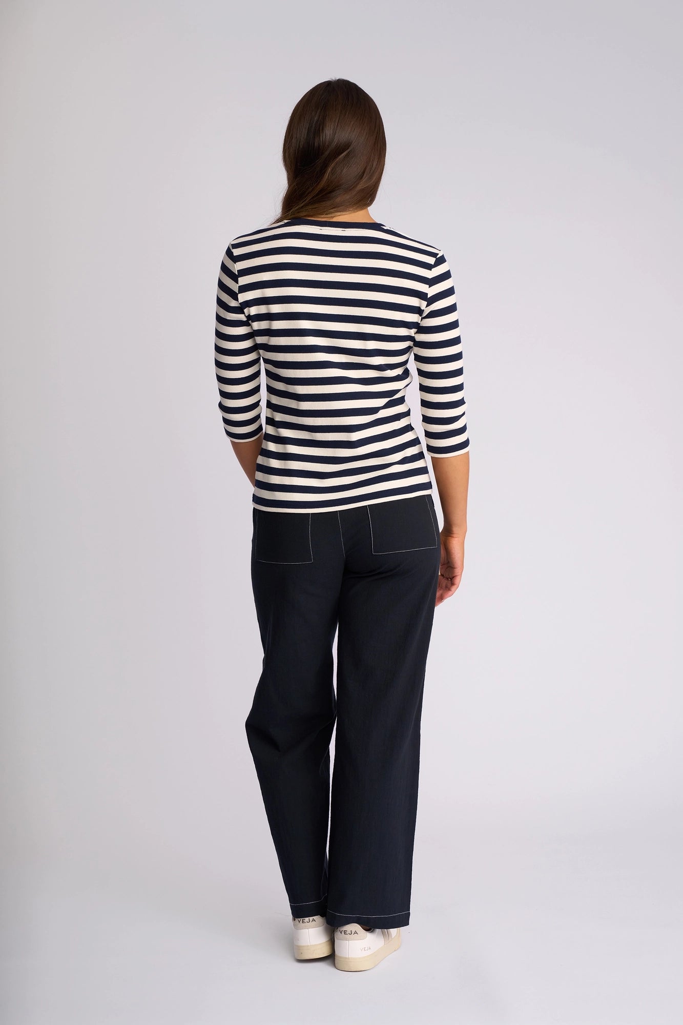 A person stands facing away, wearing the Vassalli Callie Top in Ink/Cream—a striped long-sleeve ribbed knit—paired with dark wide-leg pants featuring white stitching, and white sneakers, against a plain light background.