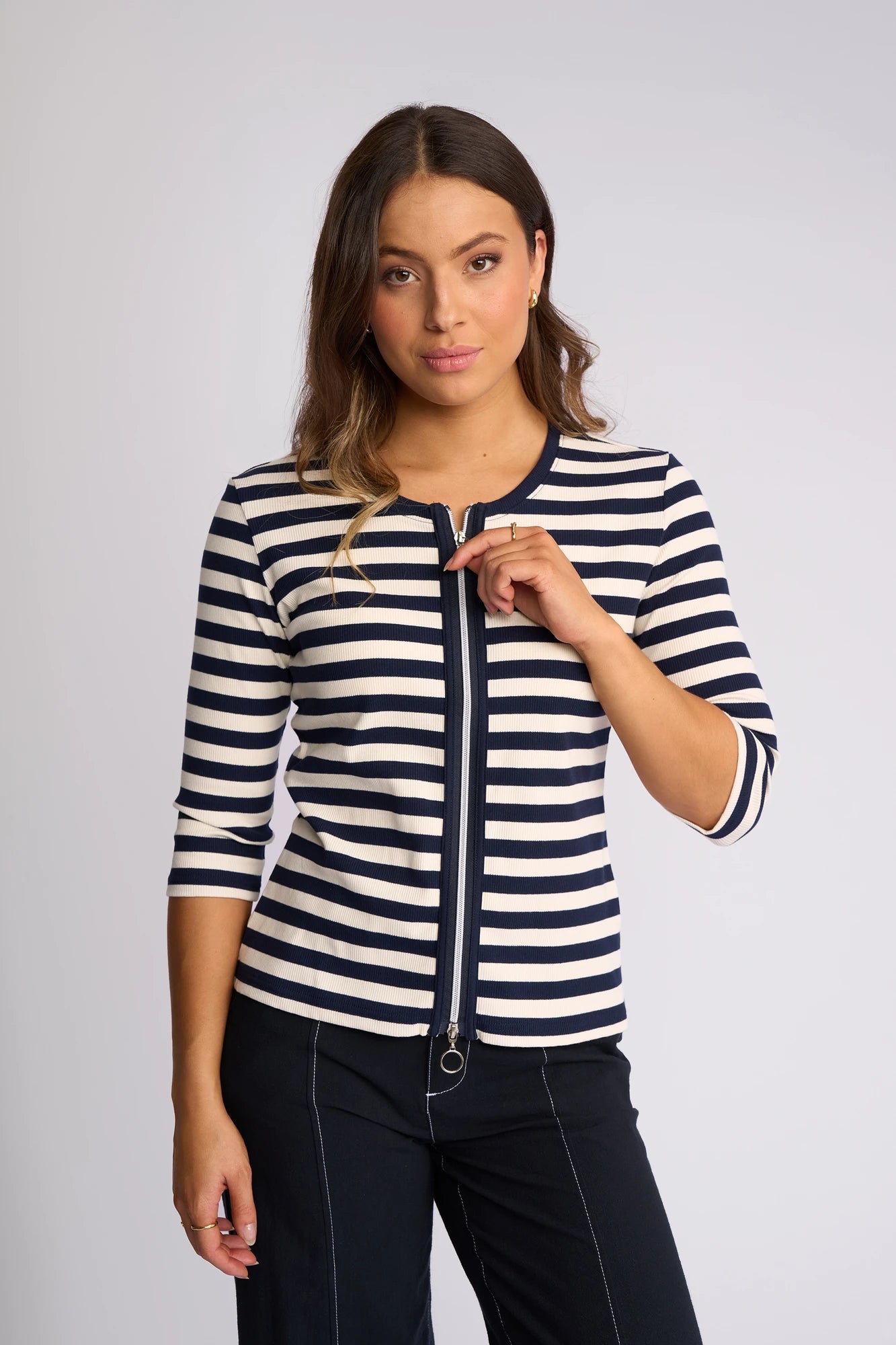 A woman with wavy brown hair wears the Vassalli Callie Top in Ink/Cream—a ribbed knit top with ink and cream stripes, a two-way zip front, and three-quarter sleeves—paired with dark pants. She stands against a light background, looking at the camera with a neutral expression.