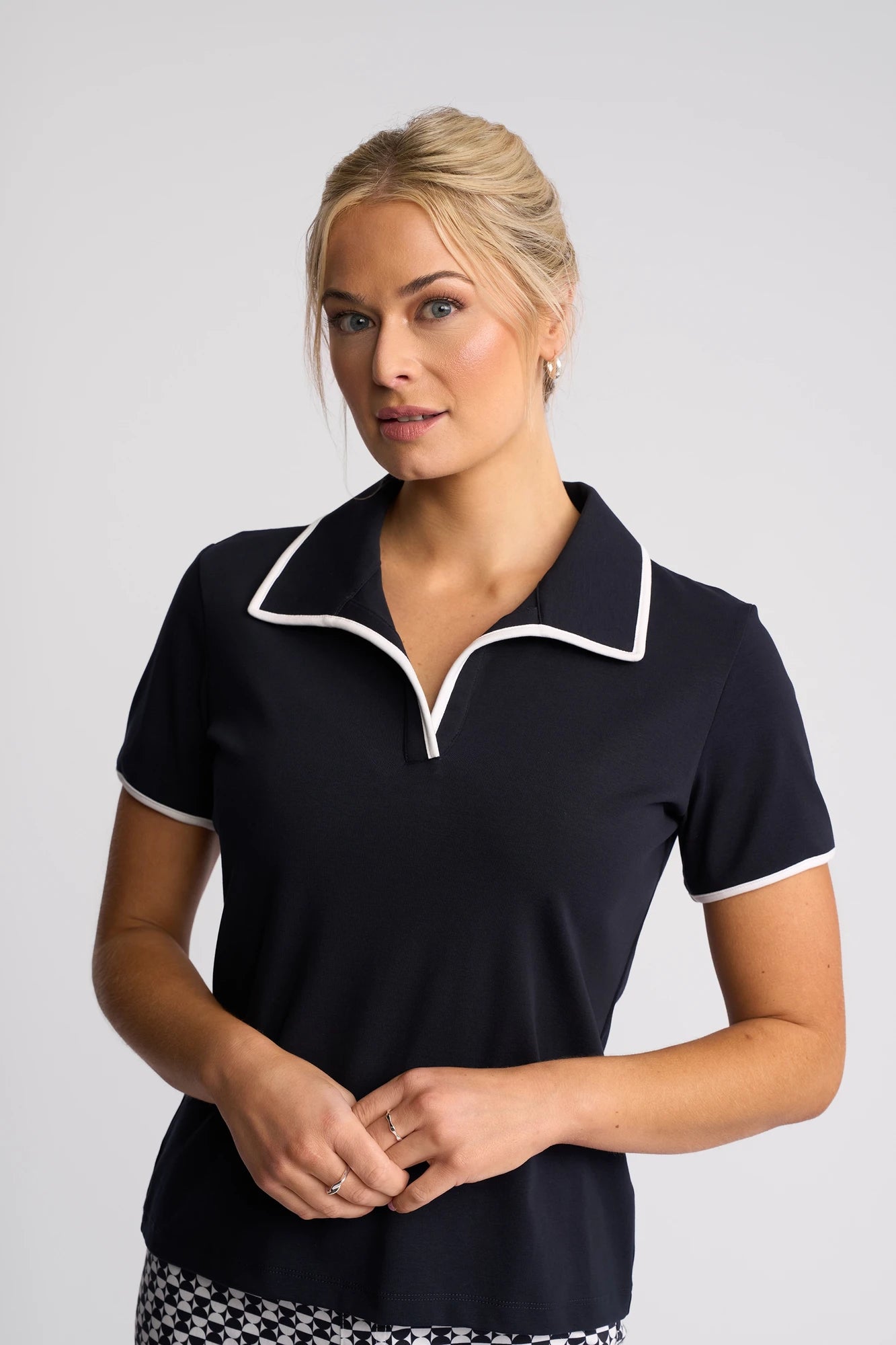 A woman with blonde hair in a bun wears the Vassalli Kit Polo Top in Ink/White, featuring a cotton stretch fabric and white trim. She stands against a plain light background, gazing softly to the side with her hands loosely clasped.
