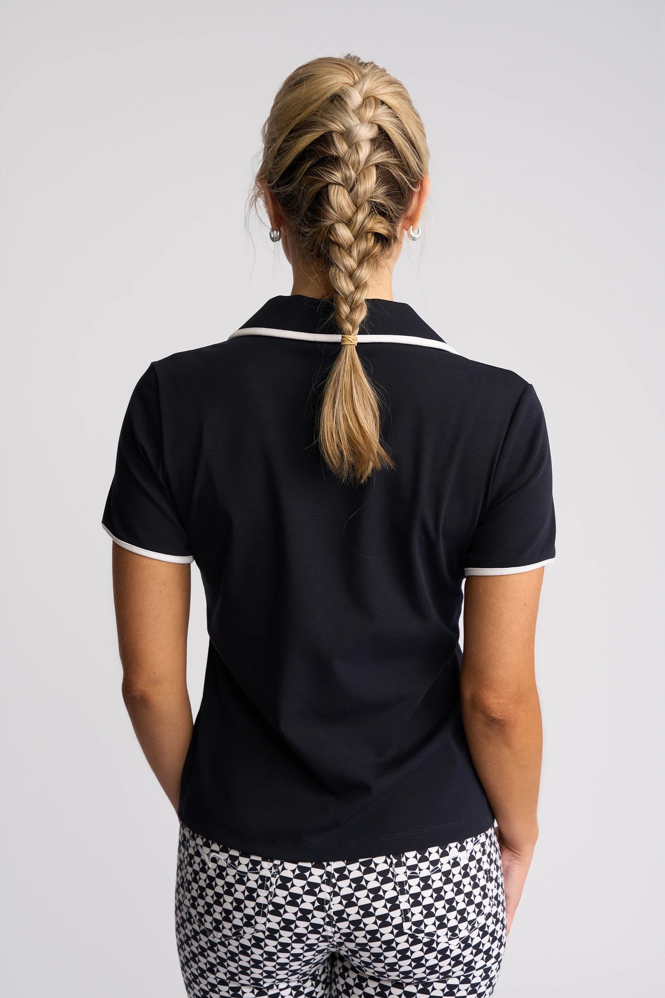 A woman with blond hair in a French braid faces away, wearing the Vassalli Kit Polo Top in Ink/White and geometric black-and-white patterned pants against a plain light background.