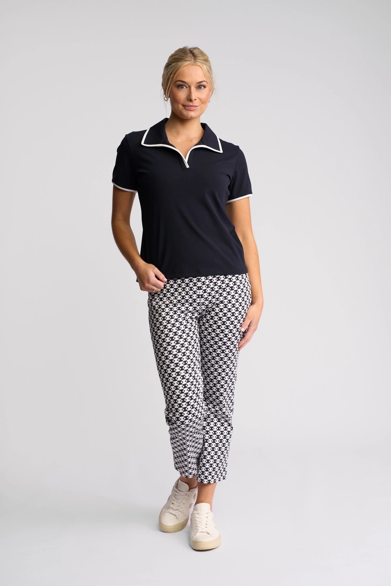 A woman stands against a plain background, smiling with one hand in her pocket. She is wearing the Vassalli Kit Polo Top in Ink/White and patterned black-and-white ankle pants crafted from cotton stretch fabric, paired with white sneakers.