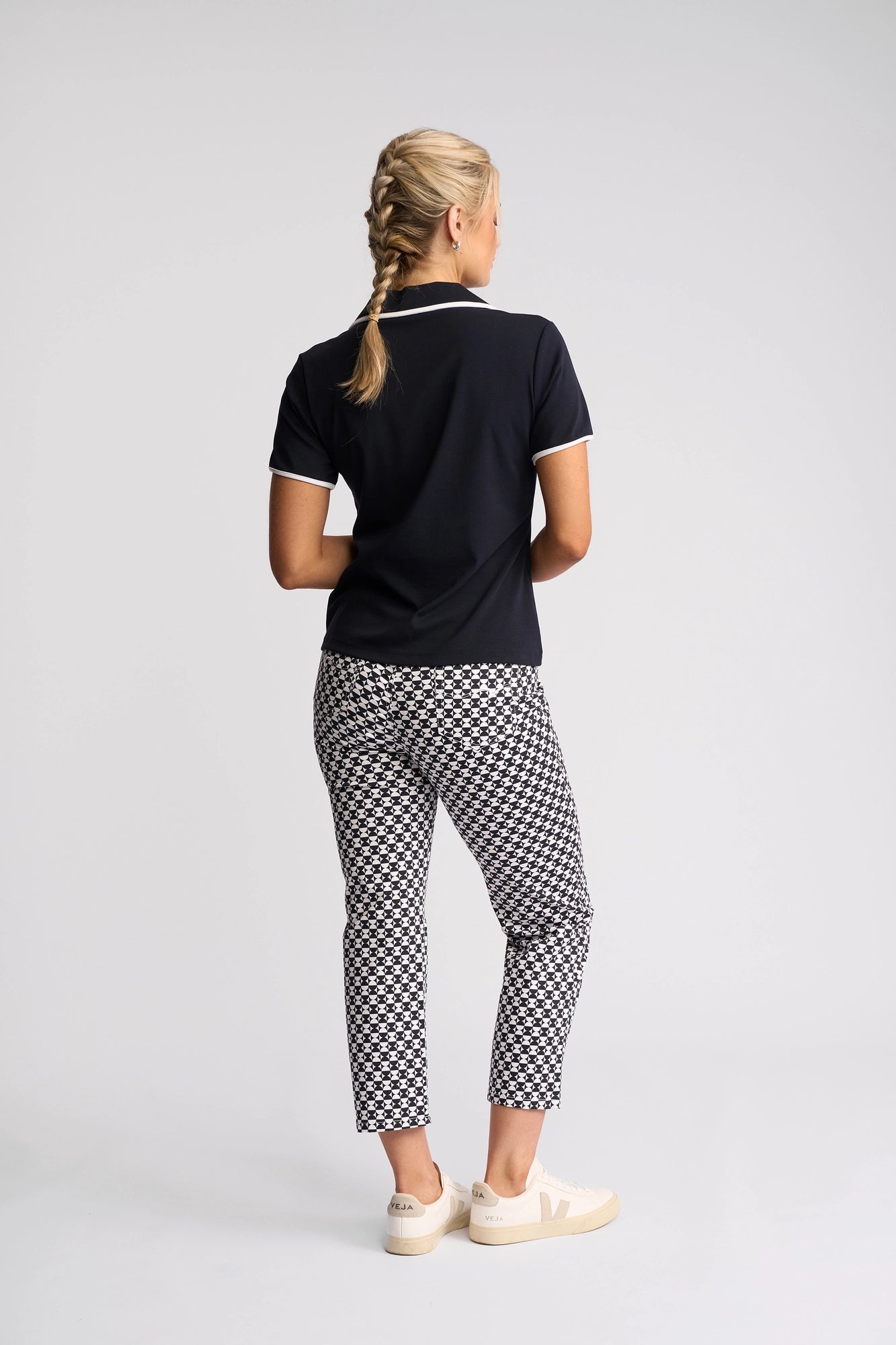 A person with blonde hair in a braid, wearing the Vassalli Kit Polo Top in Ink/White, patterned black-and-white pants, and white sneakers stands facing away from the camera against a plain background—effortlessly showcasing a sporty golf-inspired look.