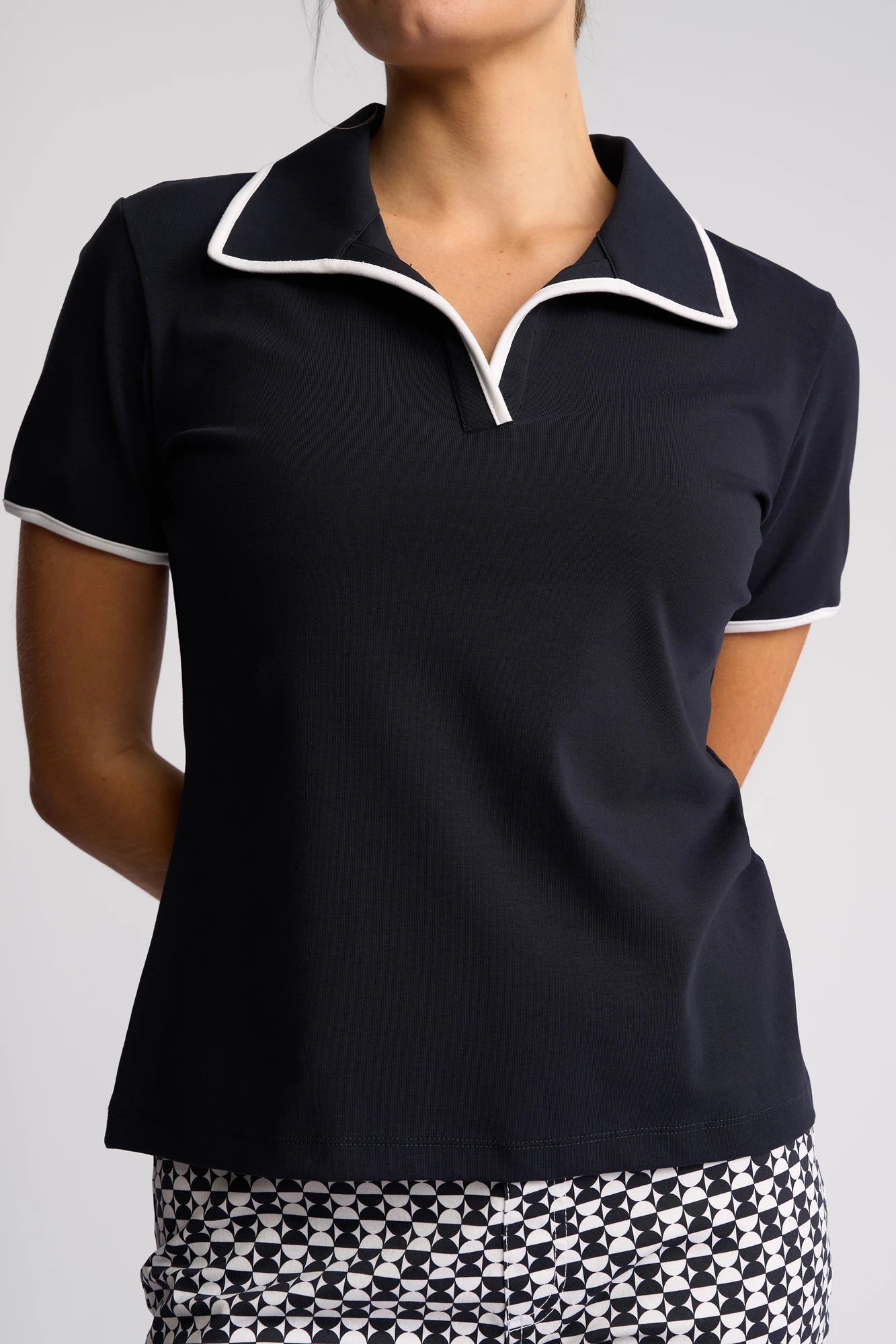 A woman stands with her arms behind her back, wearing the Vassalli Kit Polo Top in Ink/White— a black short-sleeve polo featuring white trim—paired with geometric-patterned black and white pants. Crafted from cotton stretch fabric for added comfort and flexibility, this outfit is perfect for golf or casual outings.