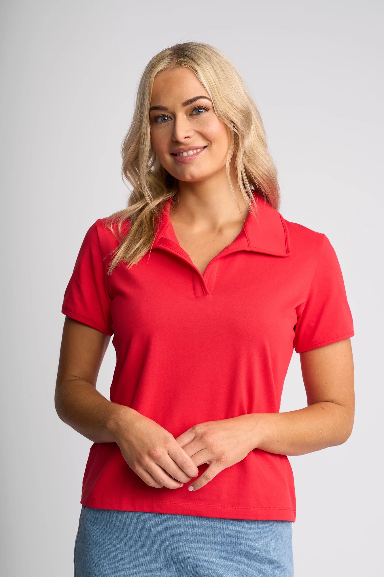 A woman with long blonde hair wears the Vassalli Kit Polo Top in Raspberry and a light blue skirt, standing against a plain light background and smiling gently at the camera.