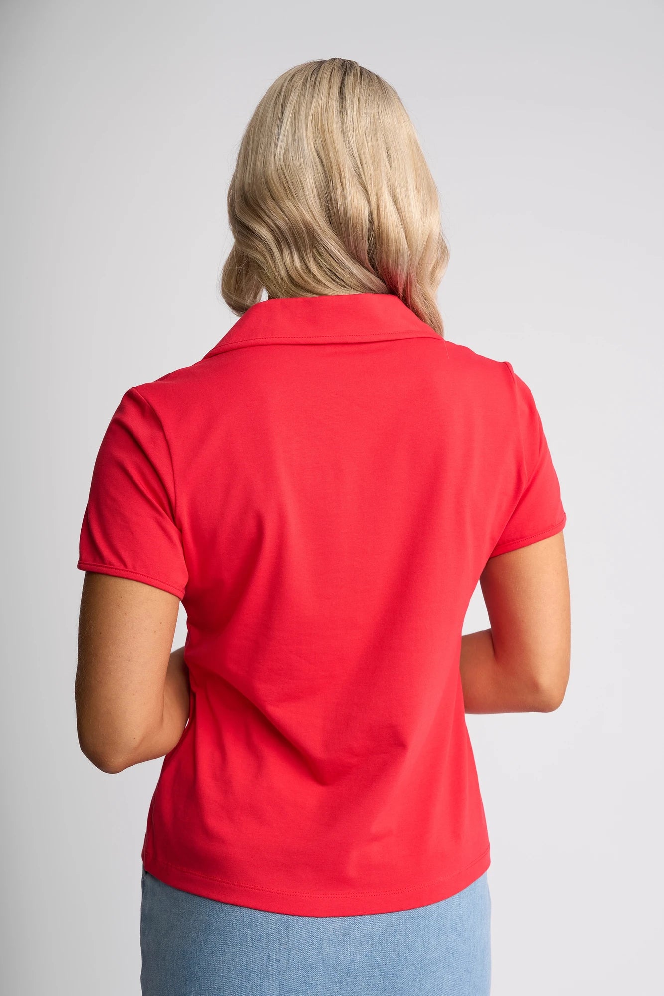 A person with blonde, wavy hair is seen from behind, wearing the Vassalli Kit Polo Top in Raspberry and a light blue skirt against a plain light background.