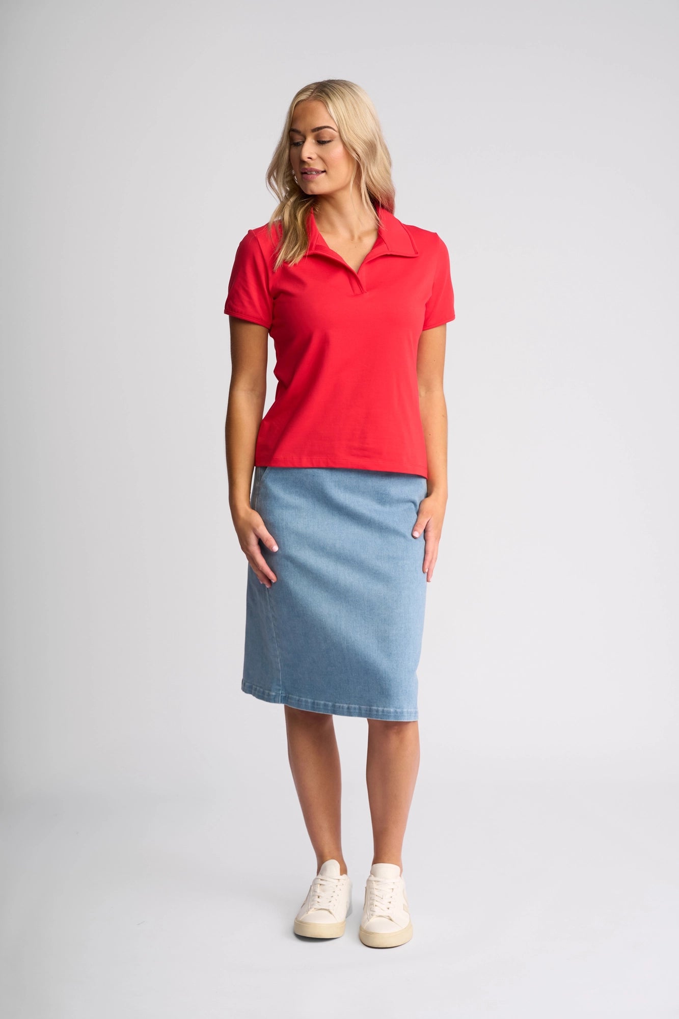 A woman stands against a plain background, wearing the Vassalli Kit Polo Top in Raspberry—a vibrant, sporty polo featuring a classic collar and short sleeves—paired with a knee-length blue denim skirt and white sneakers. She looks slightly to her left, with her hands resting by her sides.