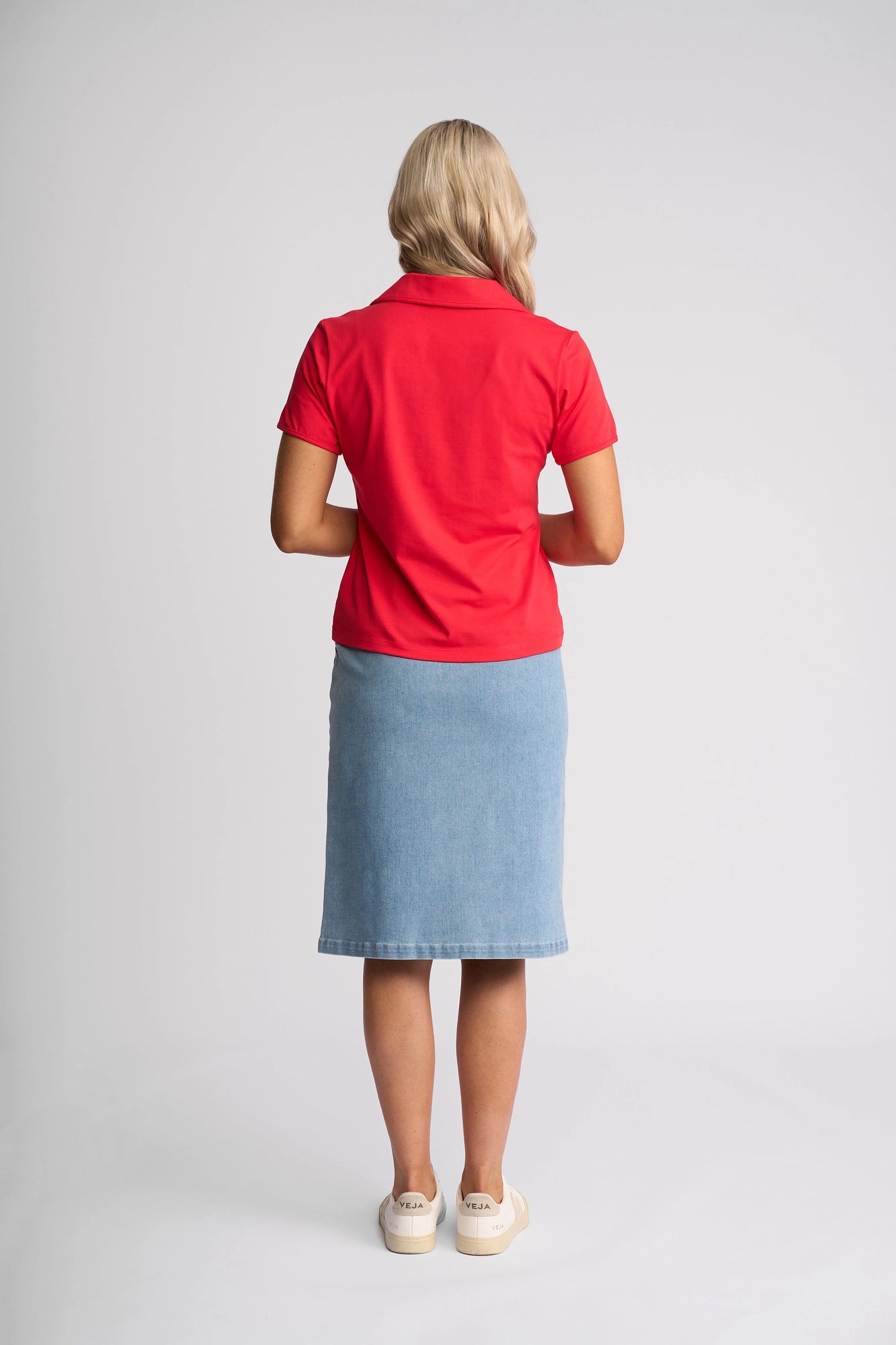 A person with blonde hair stands facing away, wearing the Vassalli Kit Polo Top in Raspberry with short sleeves, paired with a knee-length light blue denim skirt and white shoes, set against a plain light background.