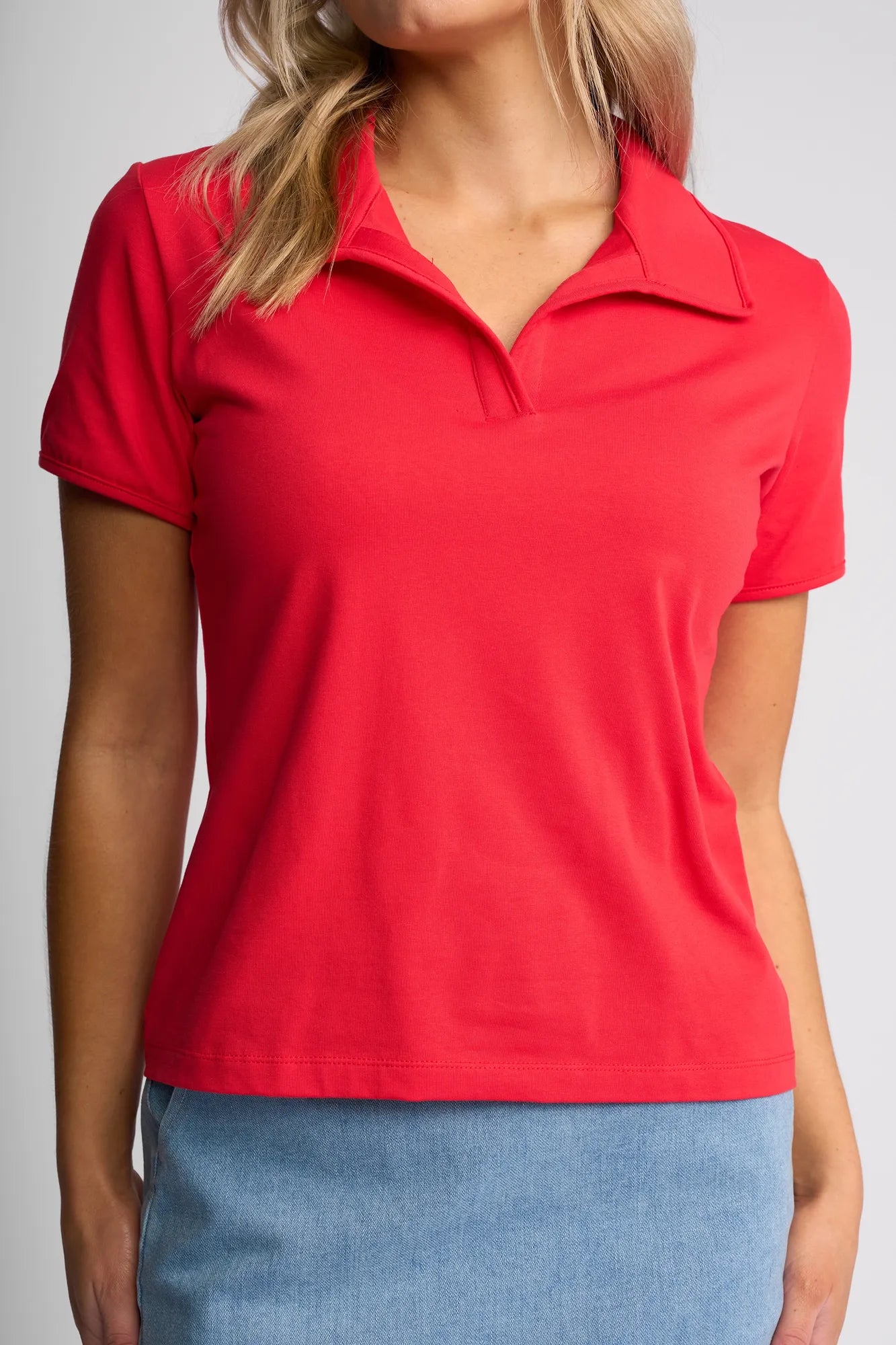 A woman wearing the Vassalli Kit Polo Top in Raspberry, a short-sleeved cotton stretch polo, paired with a light blue denim skirt, is shown from shoulders to hips standing against a plain background.
