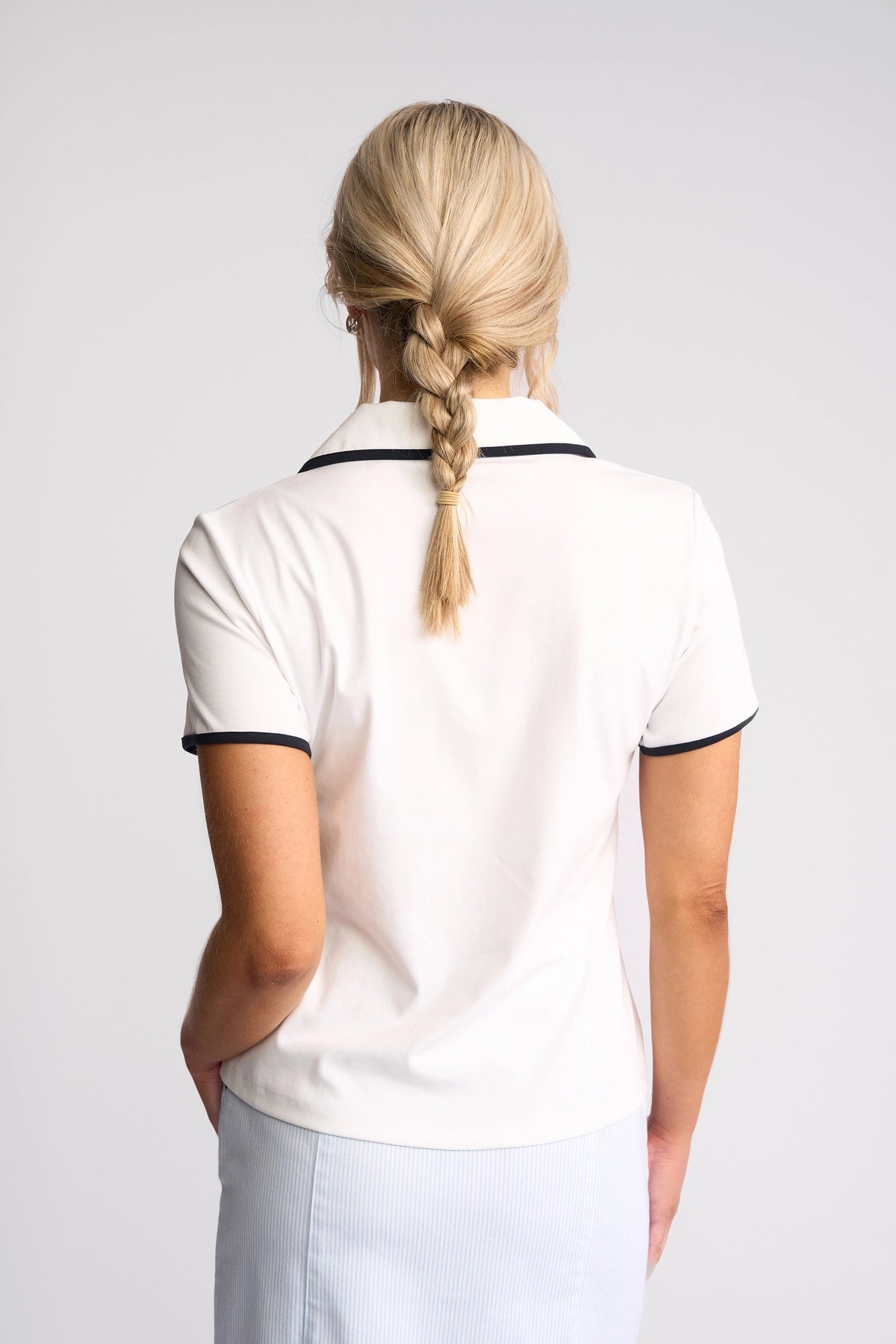 A person with blonde hair in a braid is shown from behind, wearing the Vassalli Kit Polo Top in White/Ink and a light blue skirt.