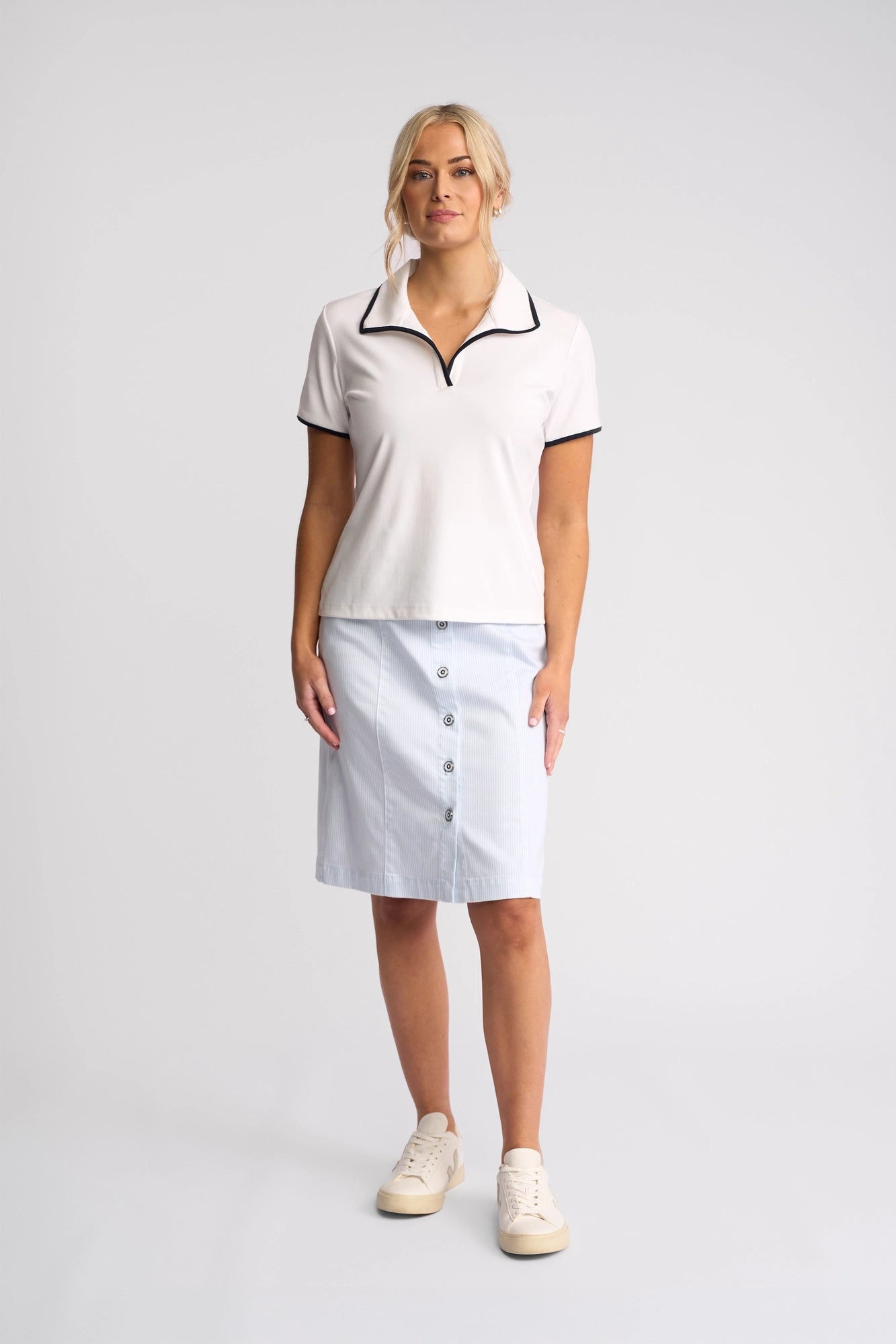 A woman stands facing forward against a plain light background, wearing the Vassalli Kit Polo Top in White/Ink—a sporty short-sleeved polo with dark trim—paired with a button-front, knee-length skirt and cream-colored sneakers.