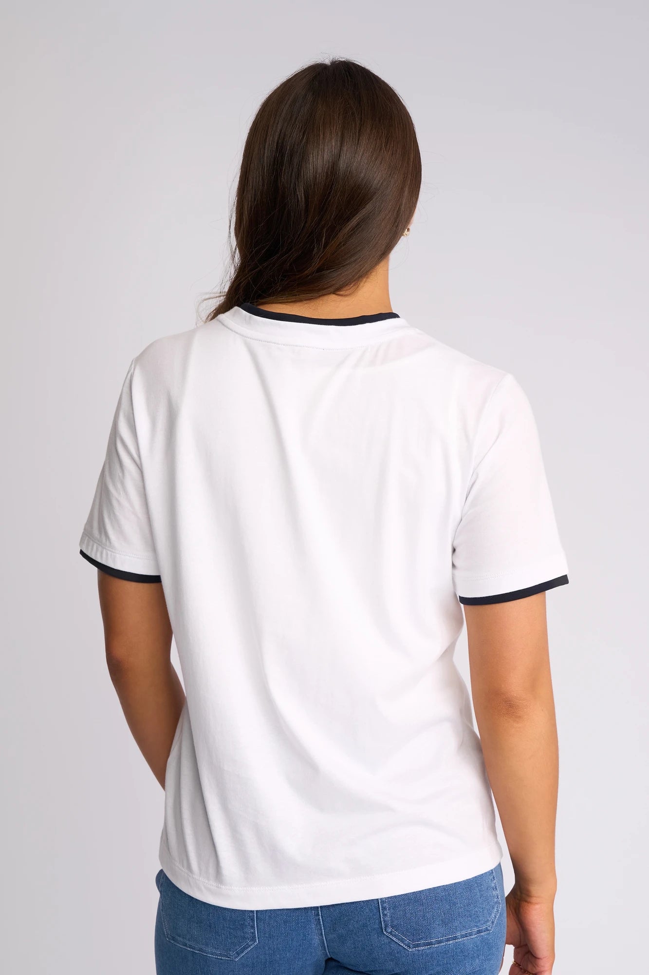 A person with long brown hair, seen from the back, is wearing a Vassalli Club Top in White/Ink and blue jeans.