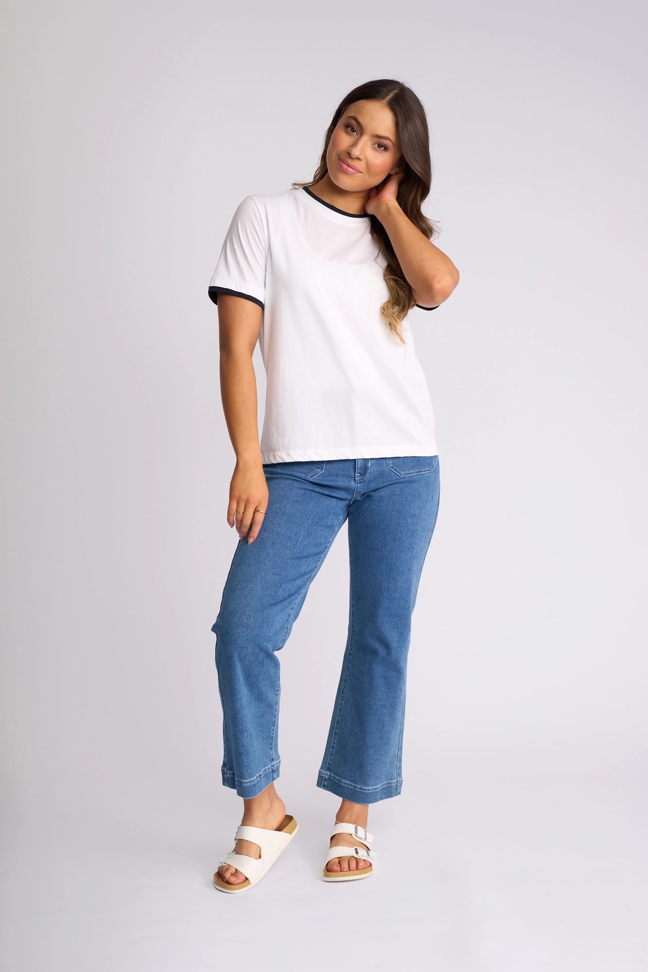 A woman stands against a plain background, touching her hair. She wears the Vassalli Club Top in White/Ink, paired with blue jeans and white sandals.
