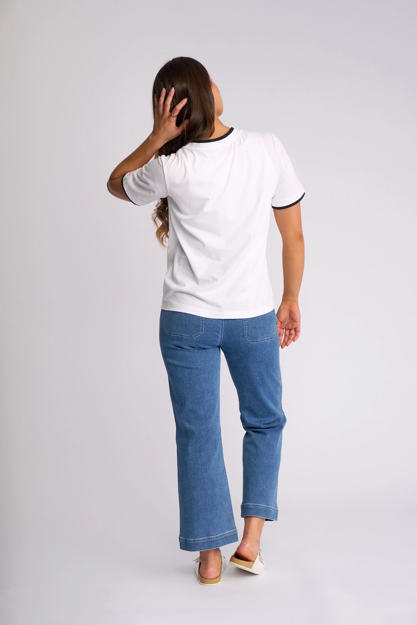 Wearing the Vassalli Club Top in White/Ink, a person with long hair, blue jeans, and light shoes stands facing away in a plain studio setting, touching their head with one hand.