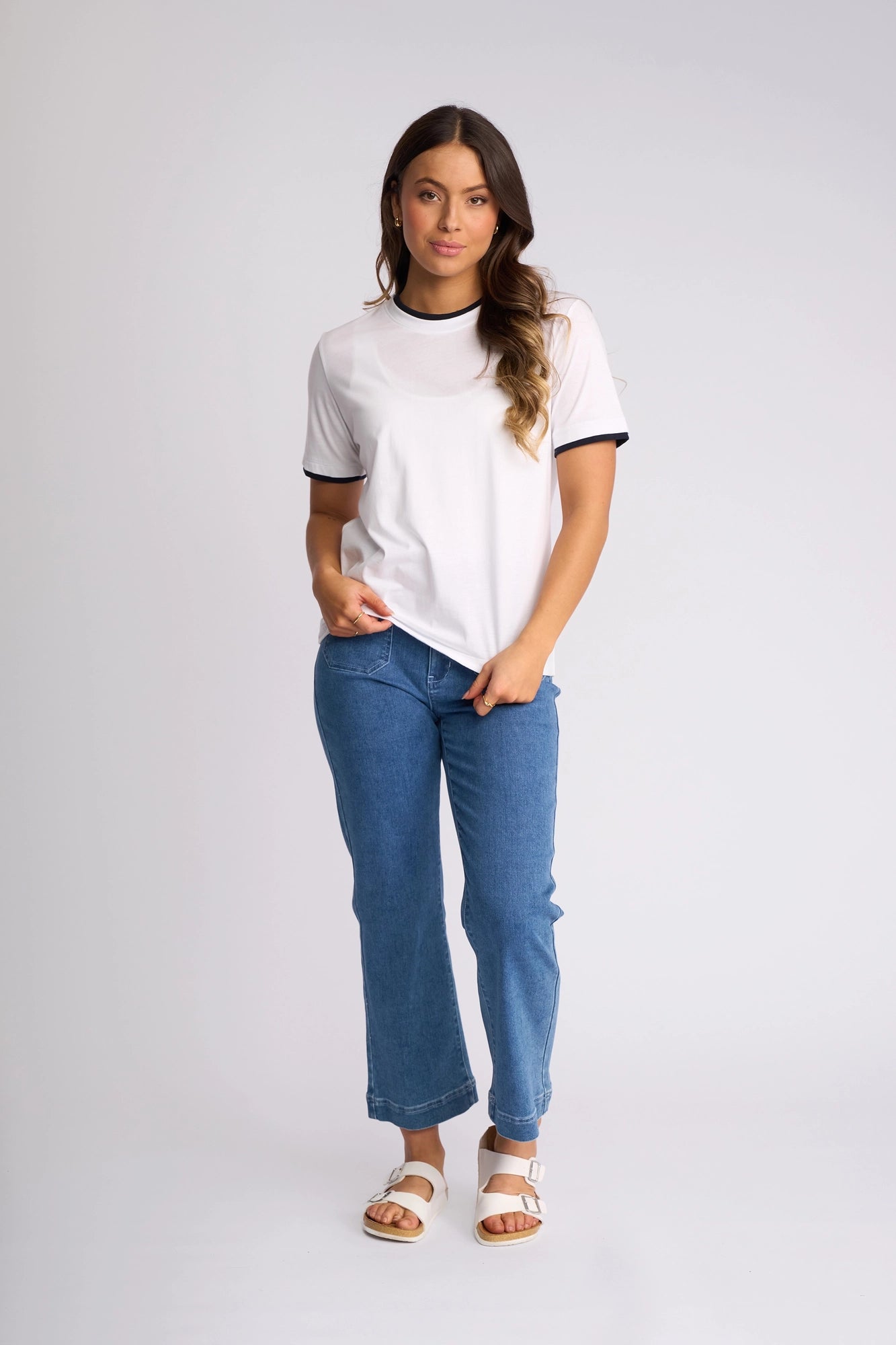 A woman with long wavy hair strikes a relaxed pose, one hand in her pocket, against a plain background. She is wearing the Vassalli Club Top in White/Ink—a sporty cotton tee with black trim—paired with blue jeans and white sandals.