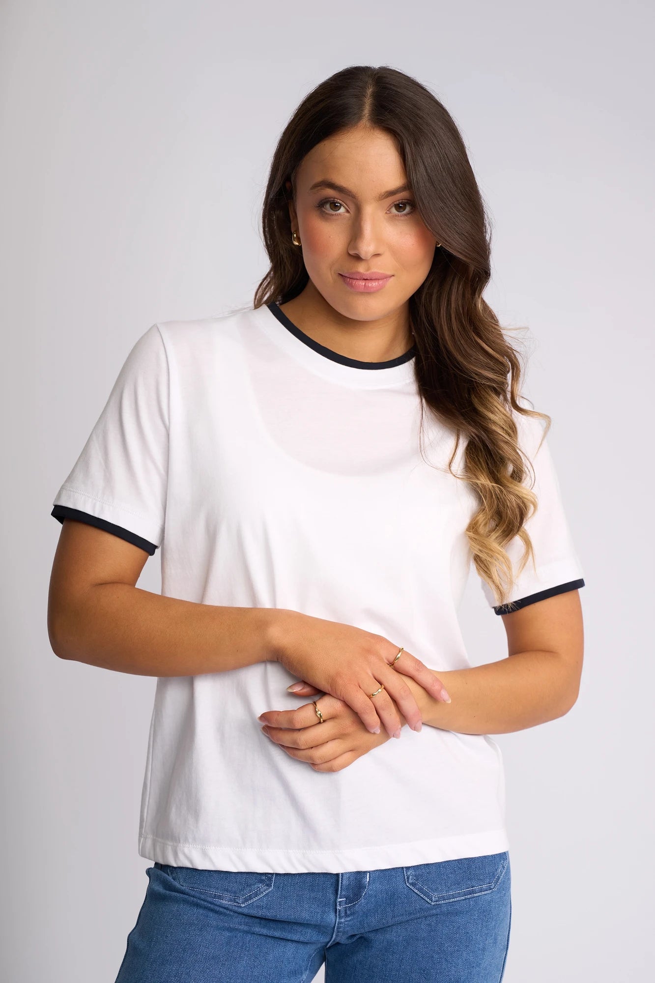 A woman with wavy brown hair wears the Vassalli Club Top in White/Ink—a sporty cotton tee featuring black trim—paired with blue jeans, as she stands against a plain light background. She looks at the camera with a slight smile, her hands gently crossed.