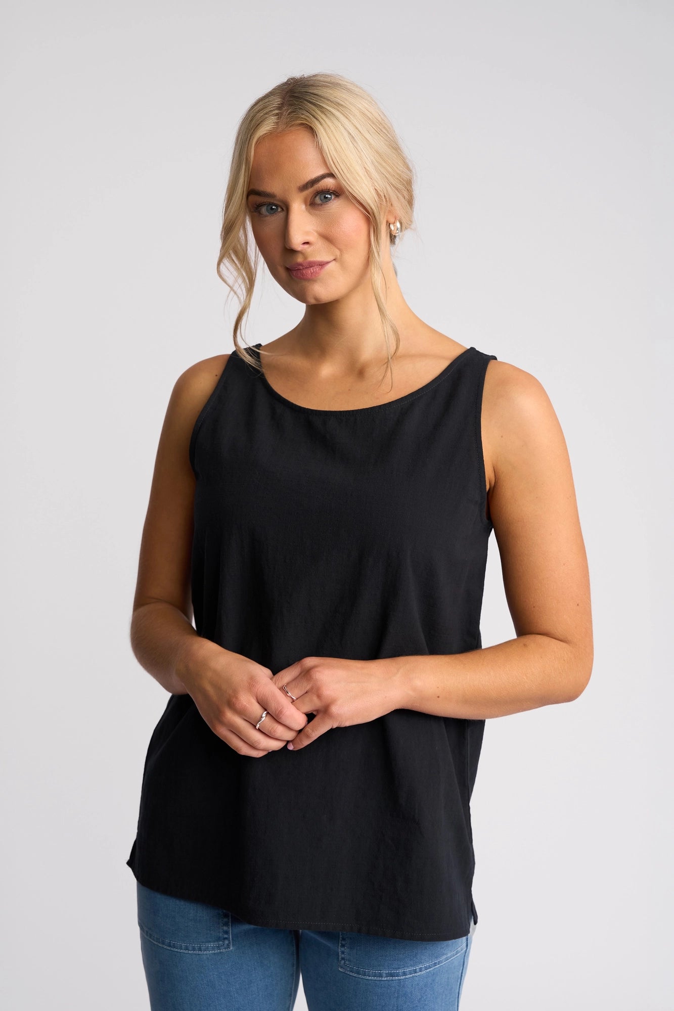 A woman with blonde hair styled in a low bun stands against a plain light background, wearing the Vassalli Romy Singlet - Ink with blue jeans. She looks at the camera with a slight smile, showcasing the lightweight cotton singlet that offers breathable, linen-look comfort for effortless style.