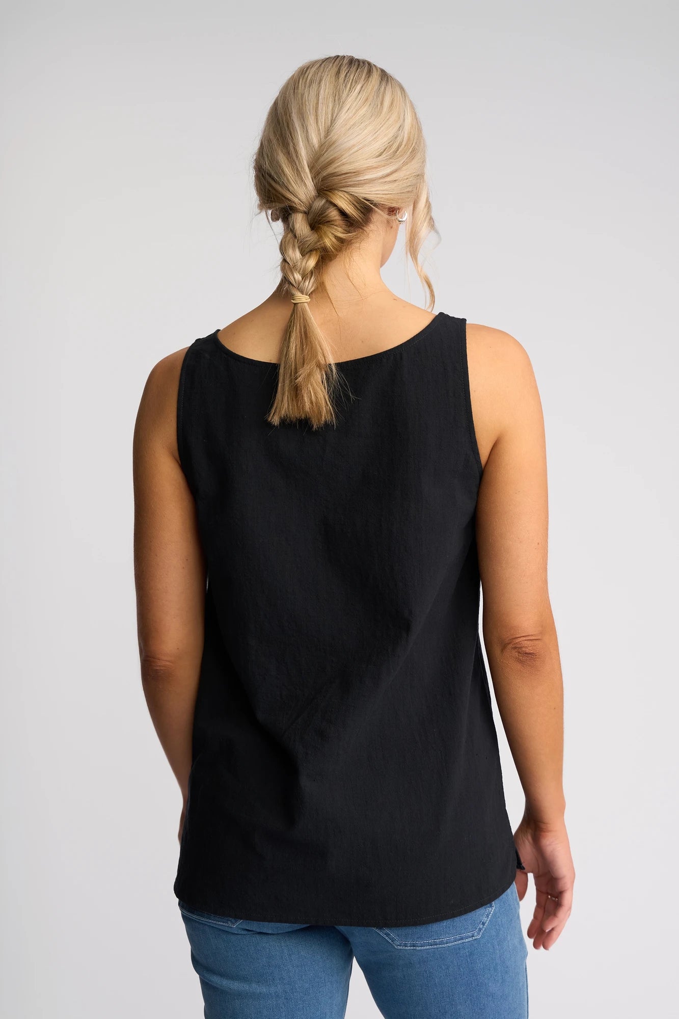 A woman with blonde hair in a braid, wearing a sleeveless black Vassalli Romy Singlet - Ink and blue jeans, stands facing away against a plain light background.