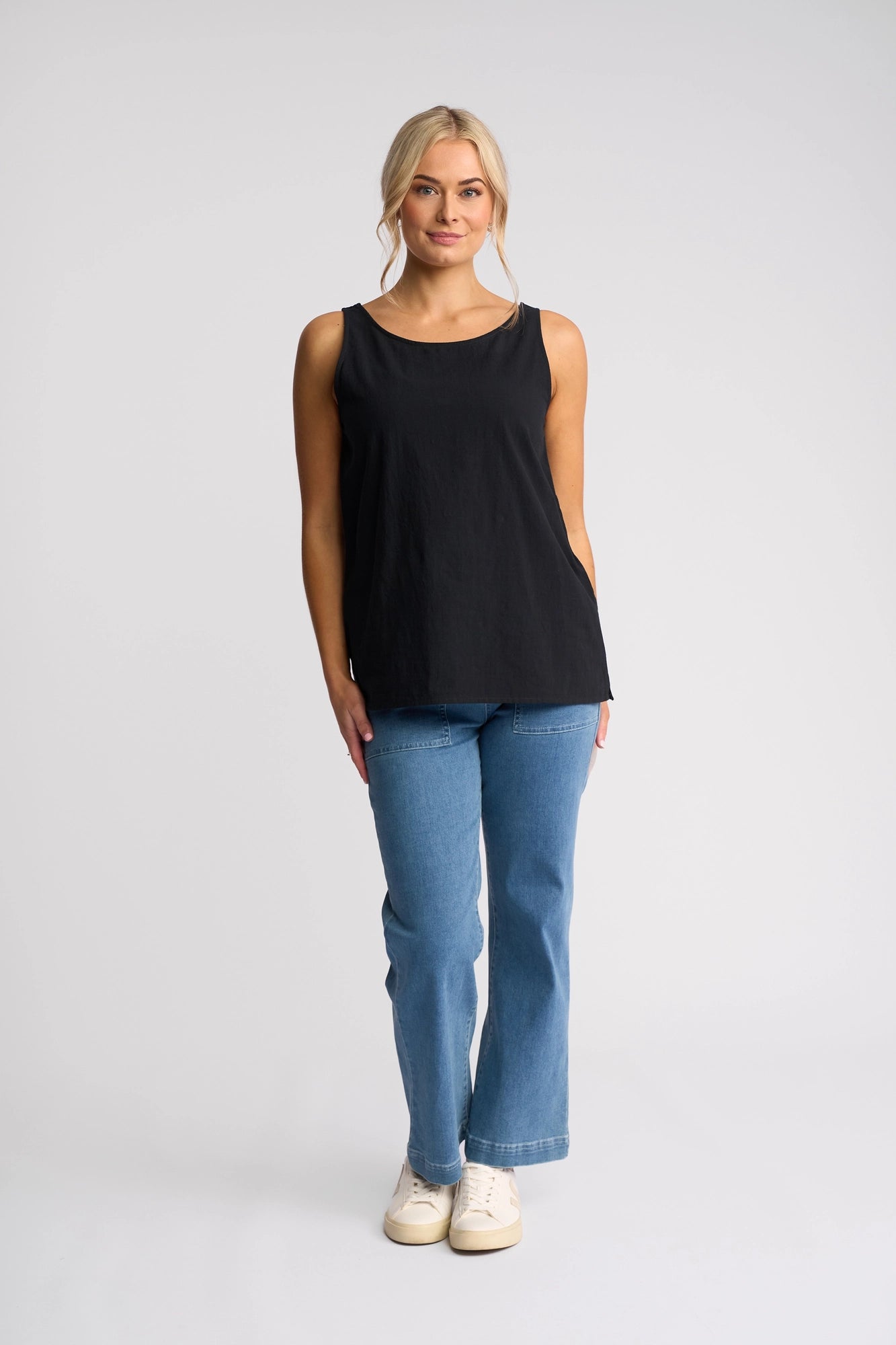 A person stands facing forward, wearing the Vassalli Romy Singlet in Ink, paired with blue jeans and white shoes, against a plain light background.