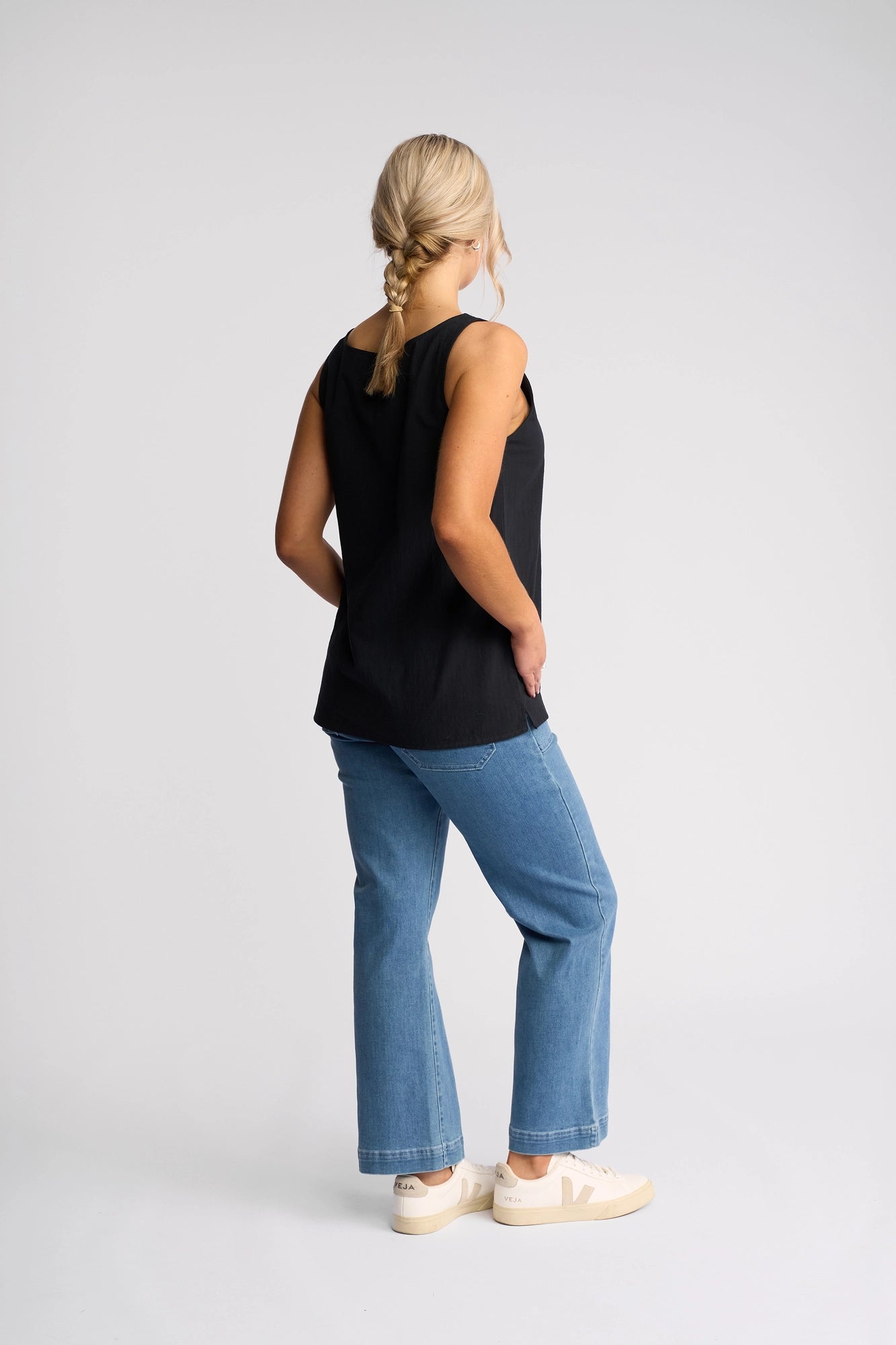 A woman with blonde hair styled in a braid, wearing the Vassalli Romy Singlet – Ink, a lightweight and stylish cotton top, paired with blue jeans and white sneakers, stands facing away against a plain light background.