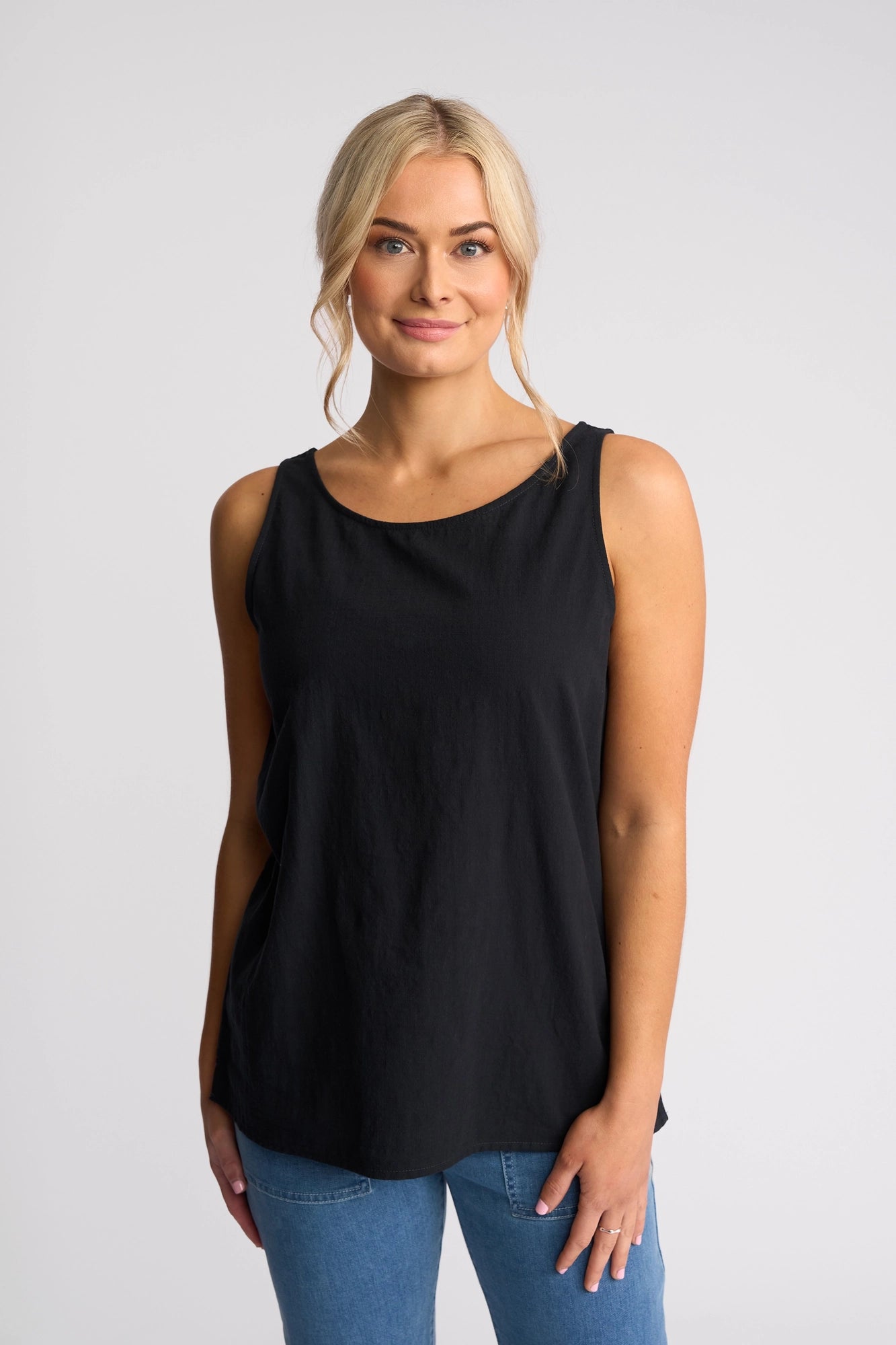 A woman with blonde hair styled in a low bun wears the Vassalli Romy Singlet - Ink, a sleeveless, lightweight cotton top with a breathable linen-look design. Paired with blue jeans, she stands against a plain white background and smiles softly at the camera—showcasing this perfect choice for relaxed summer style.
