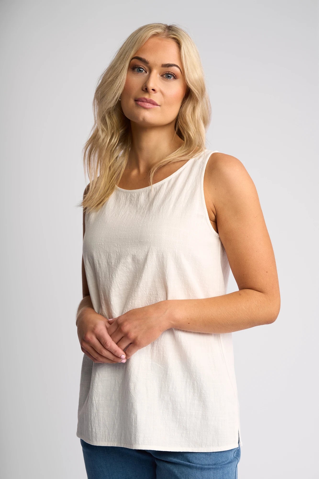 A woman with long blonde hair wears the Vassalli Romy Singlet in white, paired with blue jeans, standing against a plain light gray background and looking at the camera with a neutral expression.