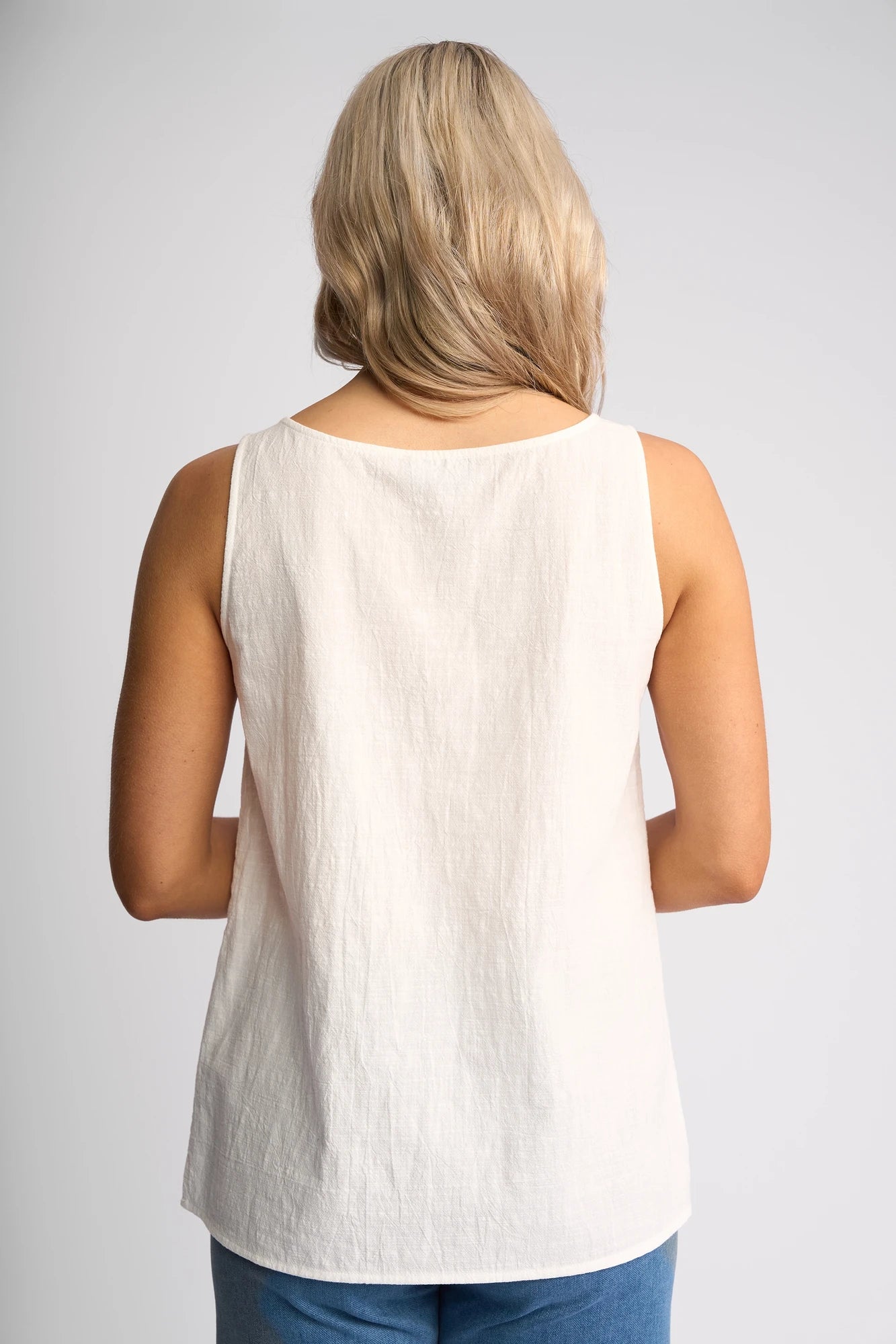 A person with light blonde hair is shown from behind against a plain light background, wearing the Vassalli Romy Singlet in white—a minimalist, sleeveless cotton top—paired with blue pants.