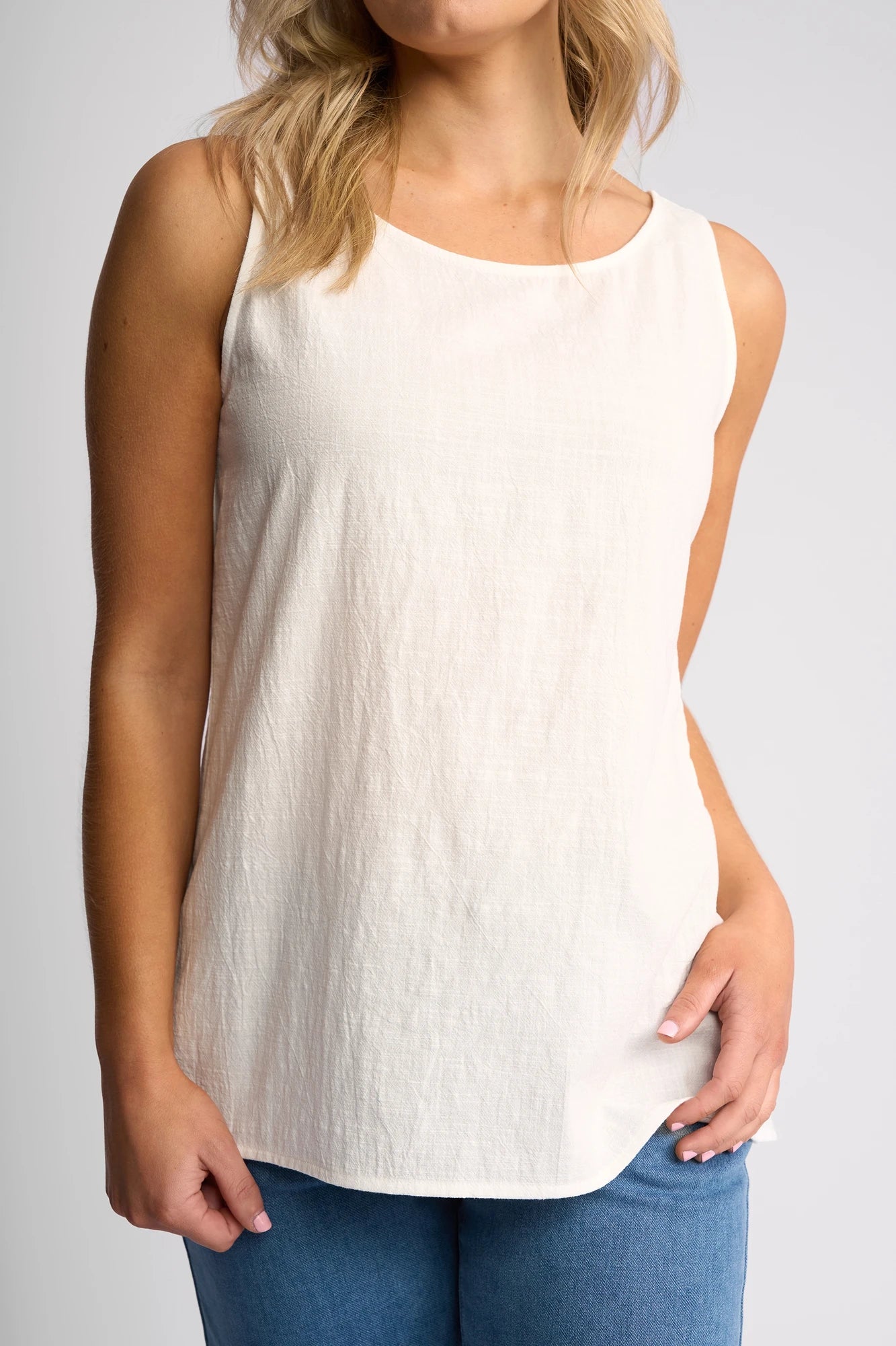 A woman with blonde hair wears the Vassalli Romy Singlet in white—a sleeveless, textured top crafted from breathable fabric—paired with blue jeans. She stands against a plain light background, one hand in her pocket and the other relaxed by her side.