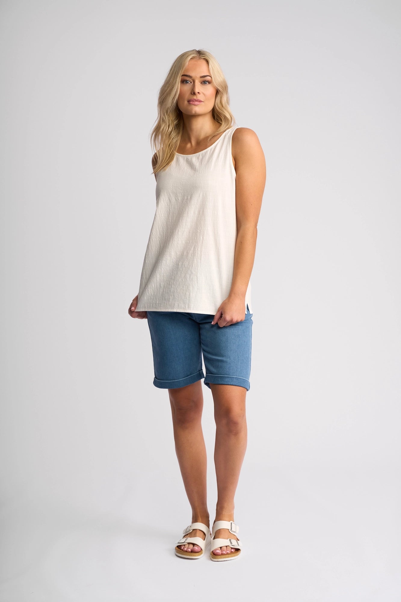 A person with long blonde hair wears the Vassalli Romy Singlet in White, paired with blue knee-length shorts and white sandals. Embracing a minimalist design, they stand facing forward against a plain light background.