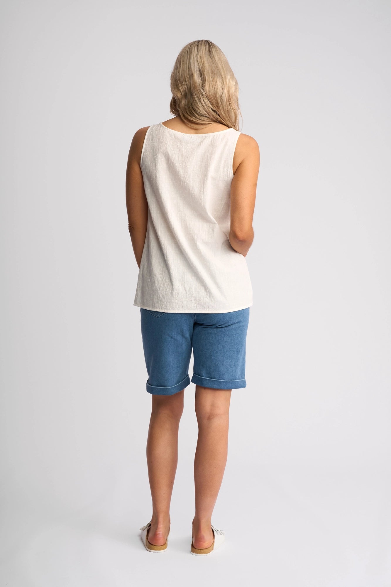 A woman with blonde hair stands facing away from the camera against a plain light background that highlights the minimalist design of the scene. She is wearing the Vassalli Romy Singlet in White—a sleeveless cotton top—paired with blue, knee-length breathable shorts and finished with white shoes for an understated look.