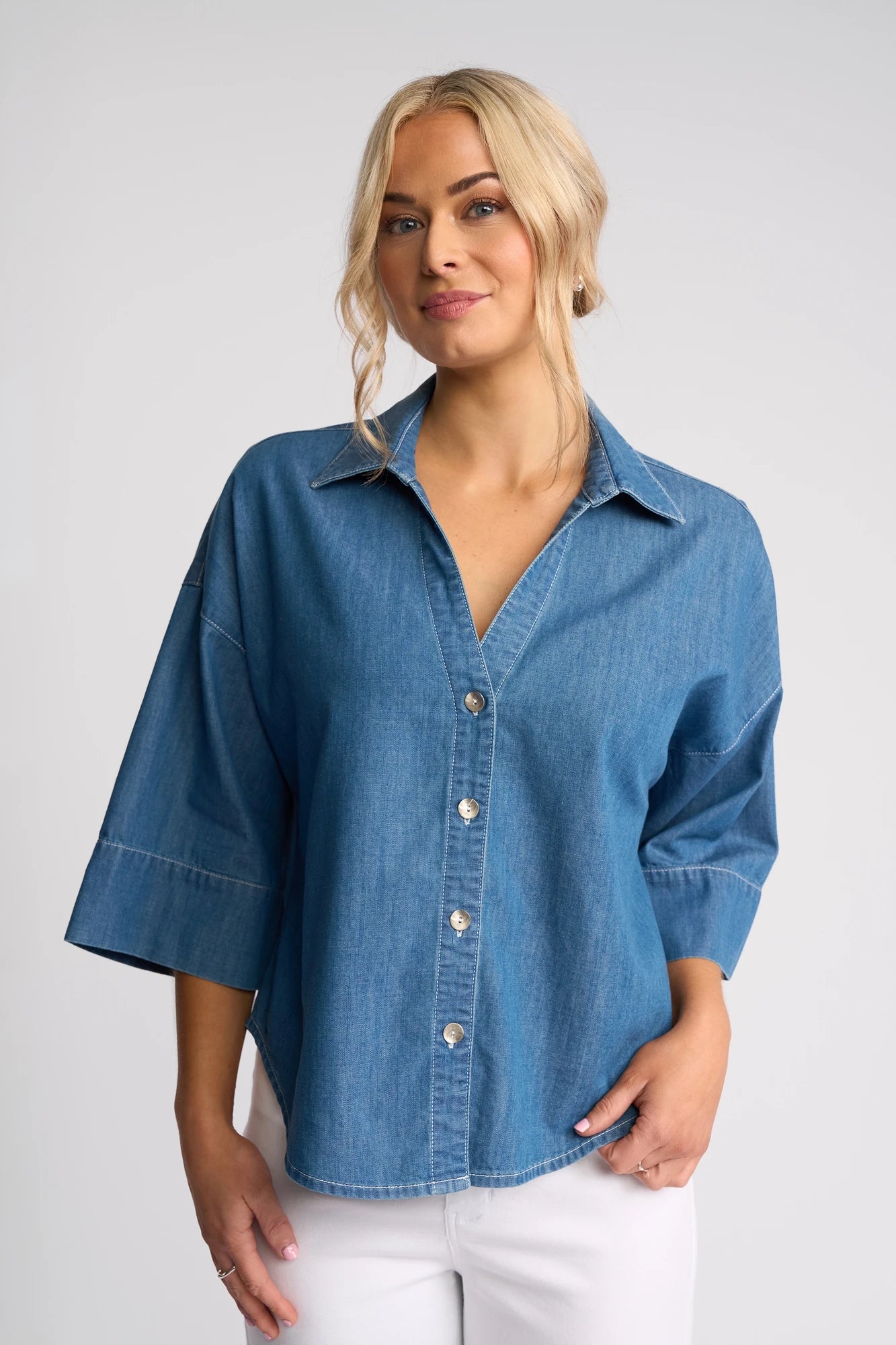 A woman with blonde hair is wearing the Vassalli Atlas Shirt in Surf Blue, a lightweight denim shirt with three-quarter sleeves, paired with white pants. She stands against a plain light background and smiles softly at the camera.